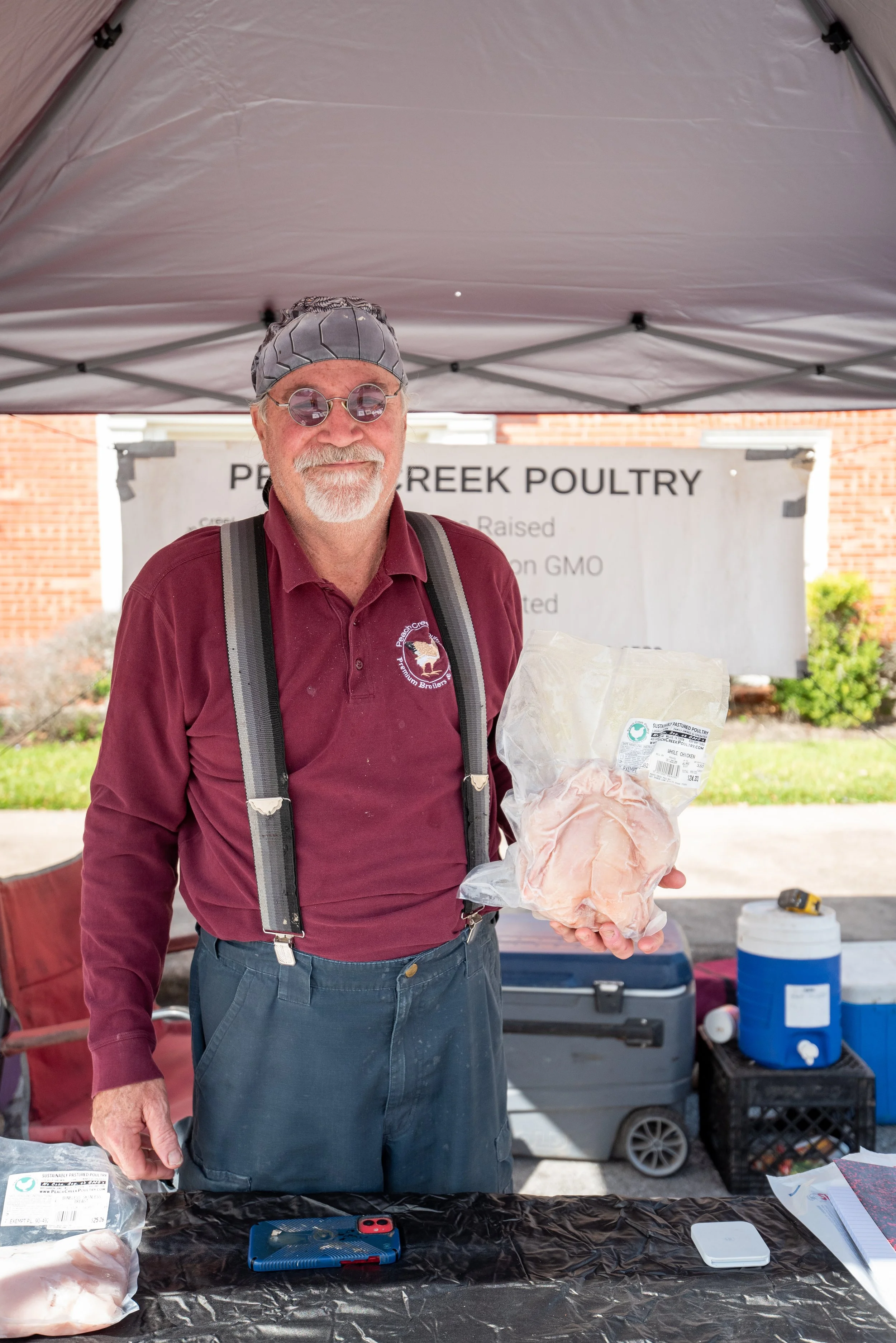 A man with a gray beard and sunglasses standing under a canopy at a farmers market, holding a packaged piece of chicken. He is wearing a maroon shirt, suspenders, and a tie-dye beanie. Background includes a banner that reads 'PEACE CREEK POULTRY' and outdoor items like coolers and a blue water jug.