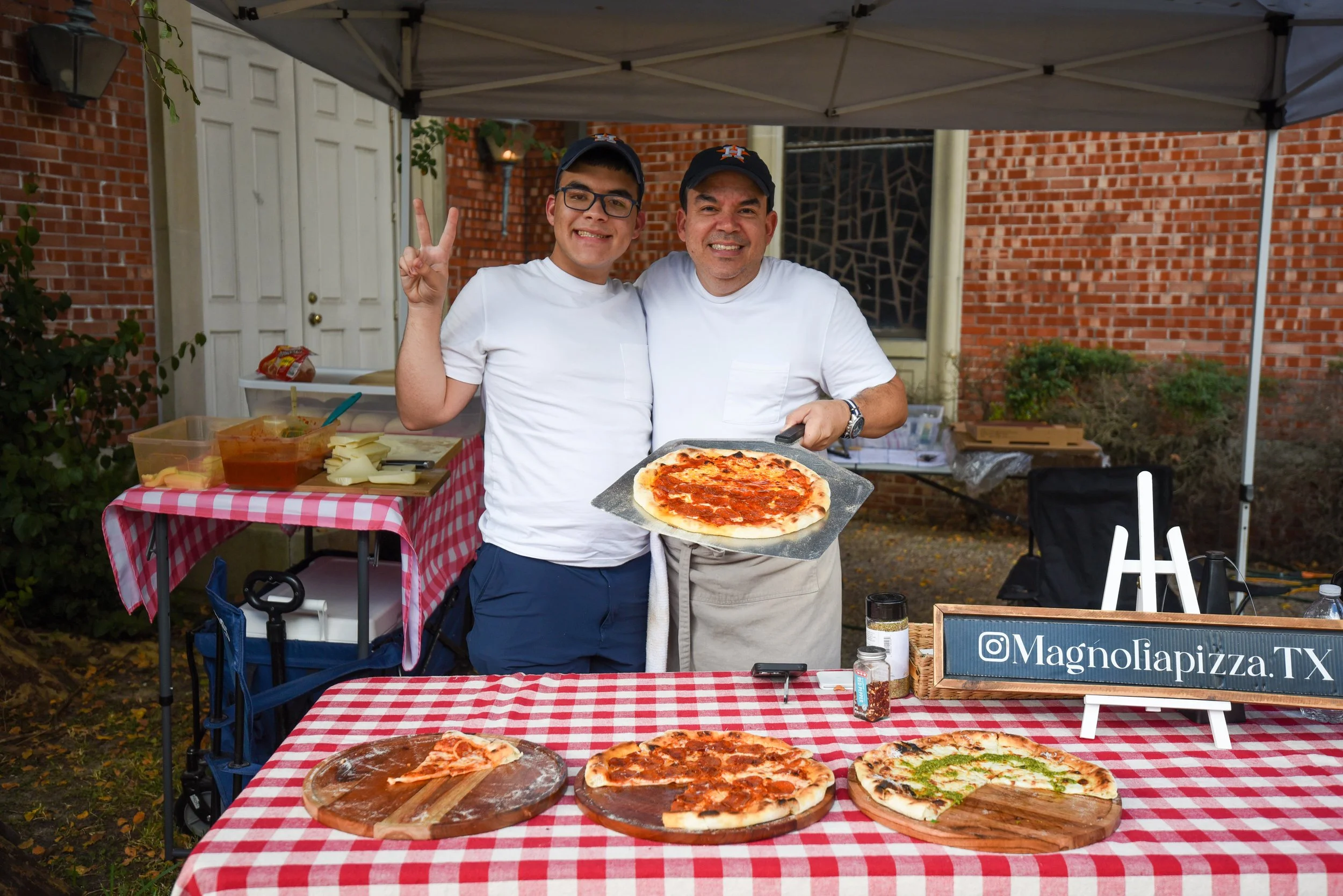 Two men standing behind a table with pizzas, one holding a pizza on a tray, smiling at the camera, under a canopy outside a building with a brick wall. The table has three pizzas, some condiments, and a sign that reads '@Magnoliapizza.TX'.