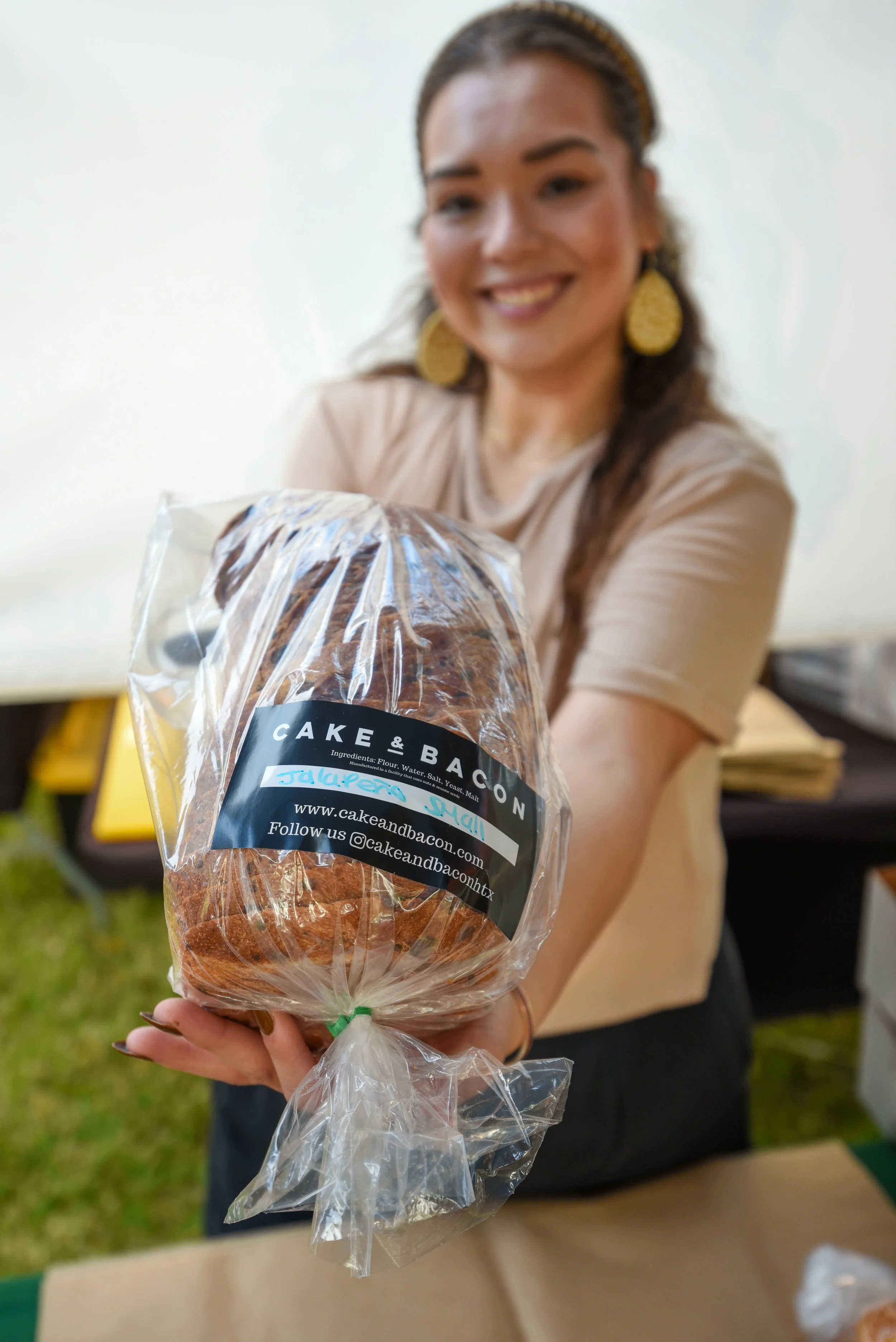 A smiling woman with long brown hair and large yellow earrings extends a bag of baked goods labeled 'Cake & Bacon' towards the camera at an outdoor event.