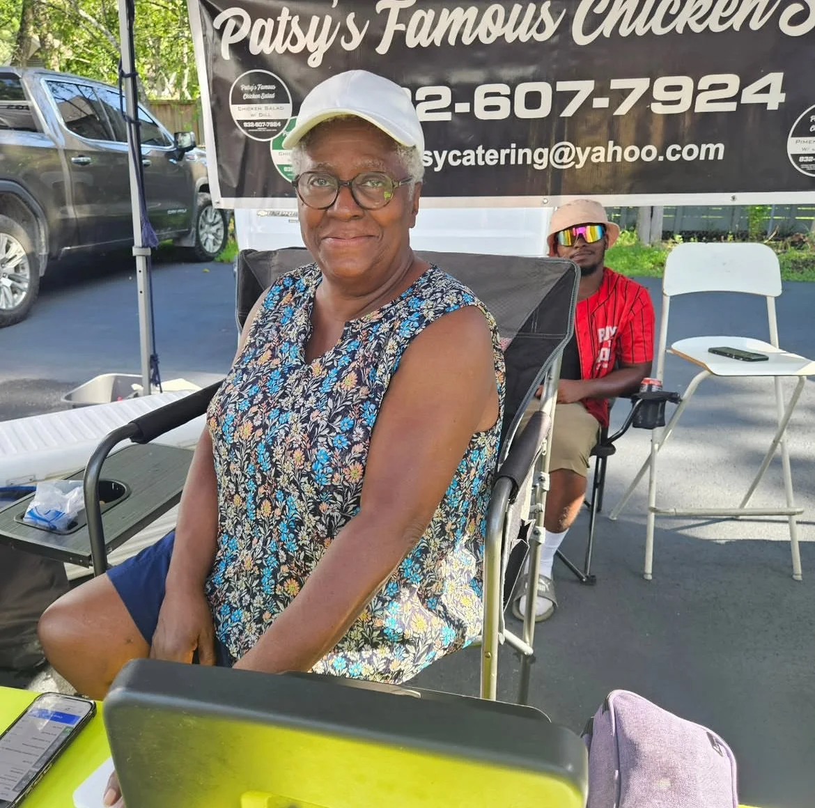 An elderly woman with gray hair, glasses, and a floral sleeveless blouse sitting outdoors at a table with a smile. Behind her, a young man wearing sunglasses, a pink cap, and a red T-shirt is seated. There is a large black banner advertising Patsy's Famous Chickens and a parking lot with cars in the background.