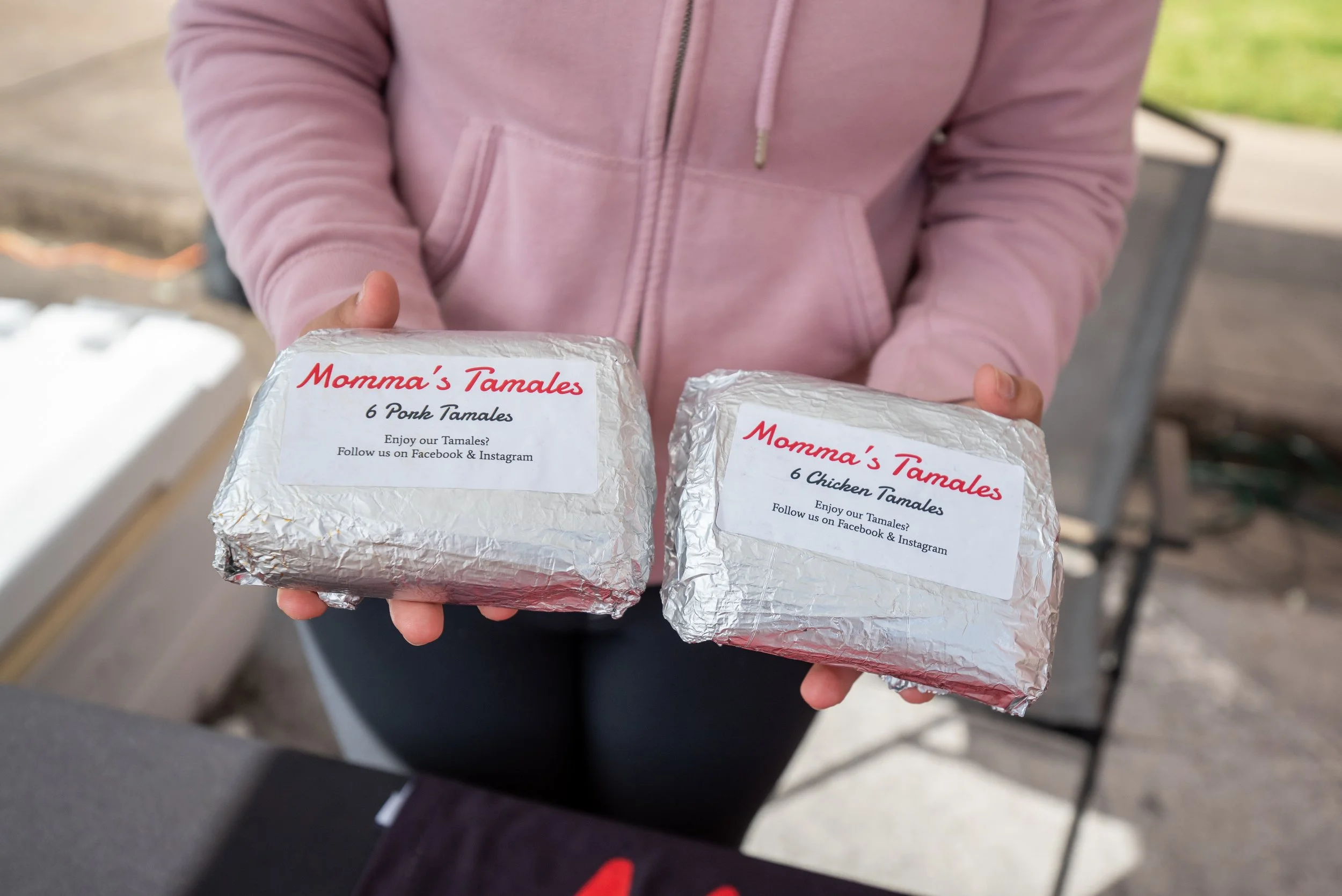 Person holding two packages of tamales wrapped in aluminum foil, labeled 'Momma's Tamales'. One package is labeled '6 Pork Tamales' and the other '6 Chicken Tamales'.