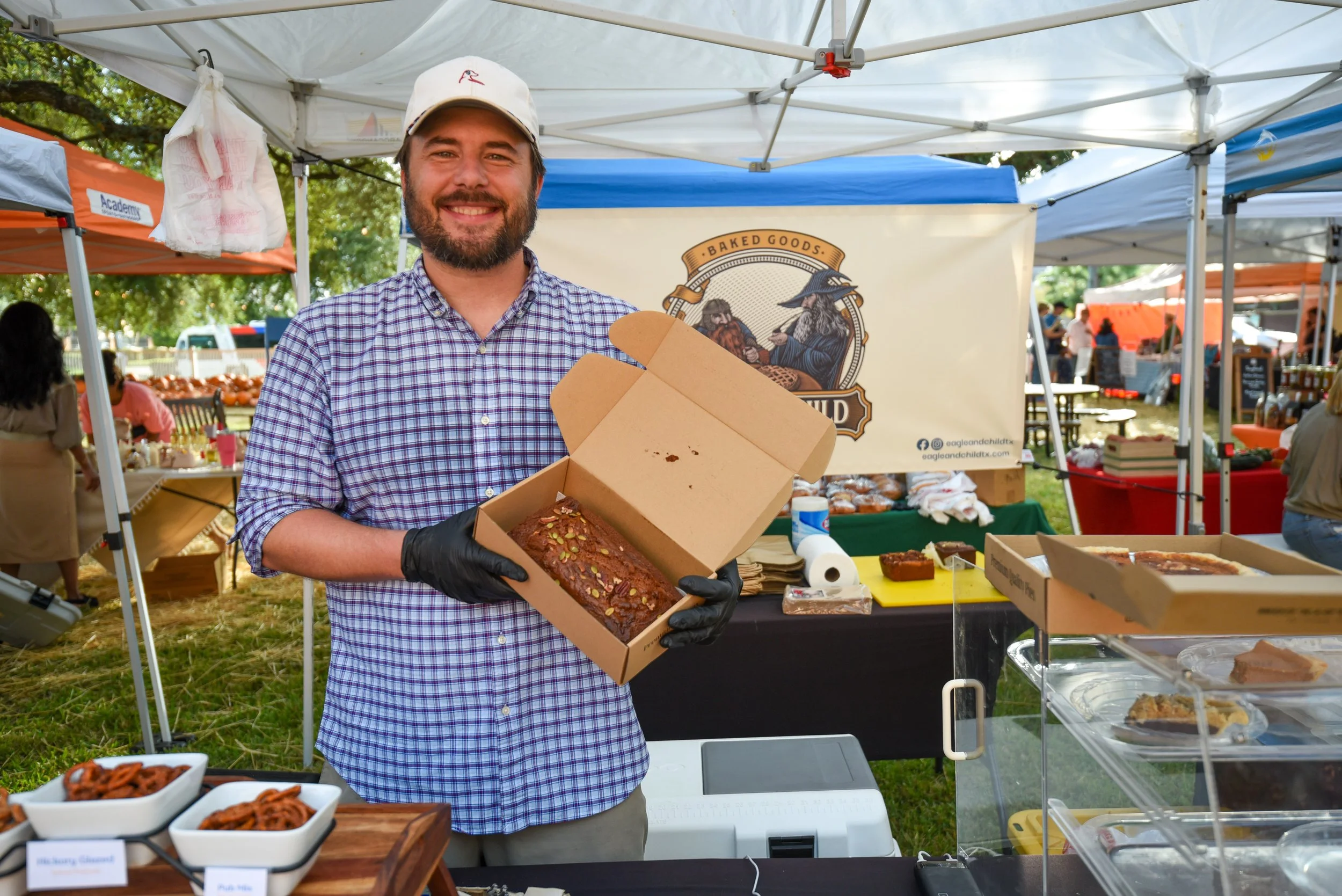 A smiling man with a beard, wearing a white cap and a checkered shirt, holding a box of baked goods at an outdoor market stand under a white canopy.