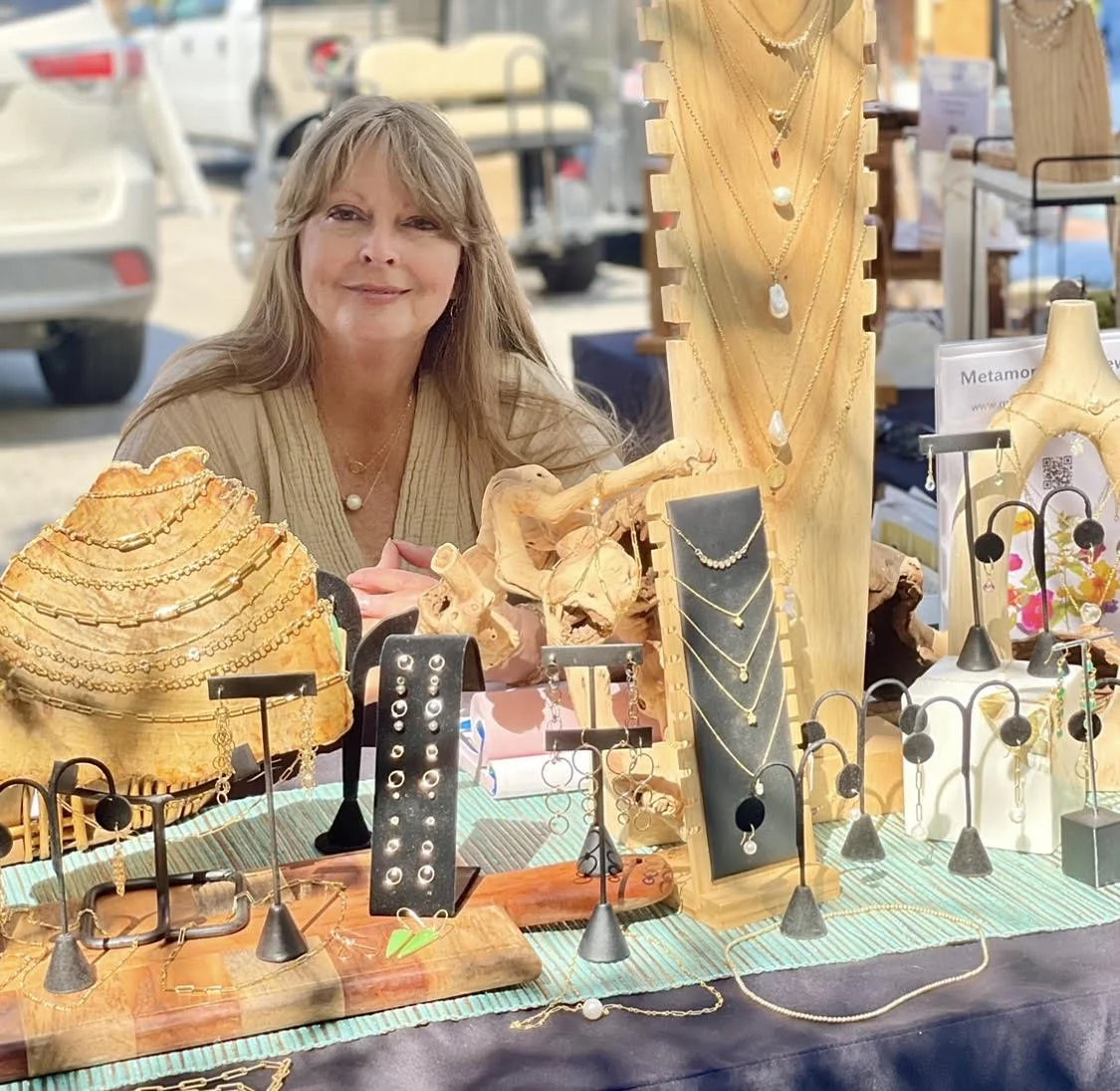 A woman sitting behind a jewelry booth at an outdoor market, with jewelry displayed on wooden stands, necklaces, bracelets, and earrings, under a white canopy, with parked cars and other market stalls in the background.