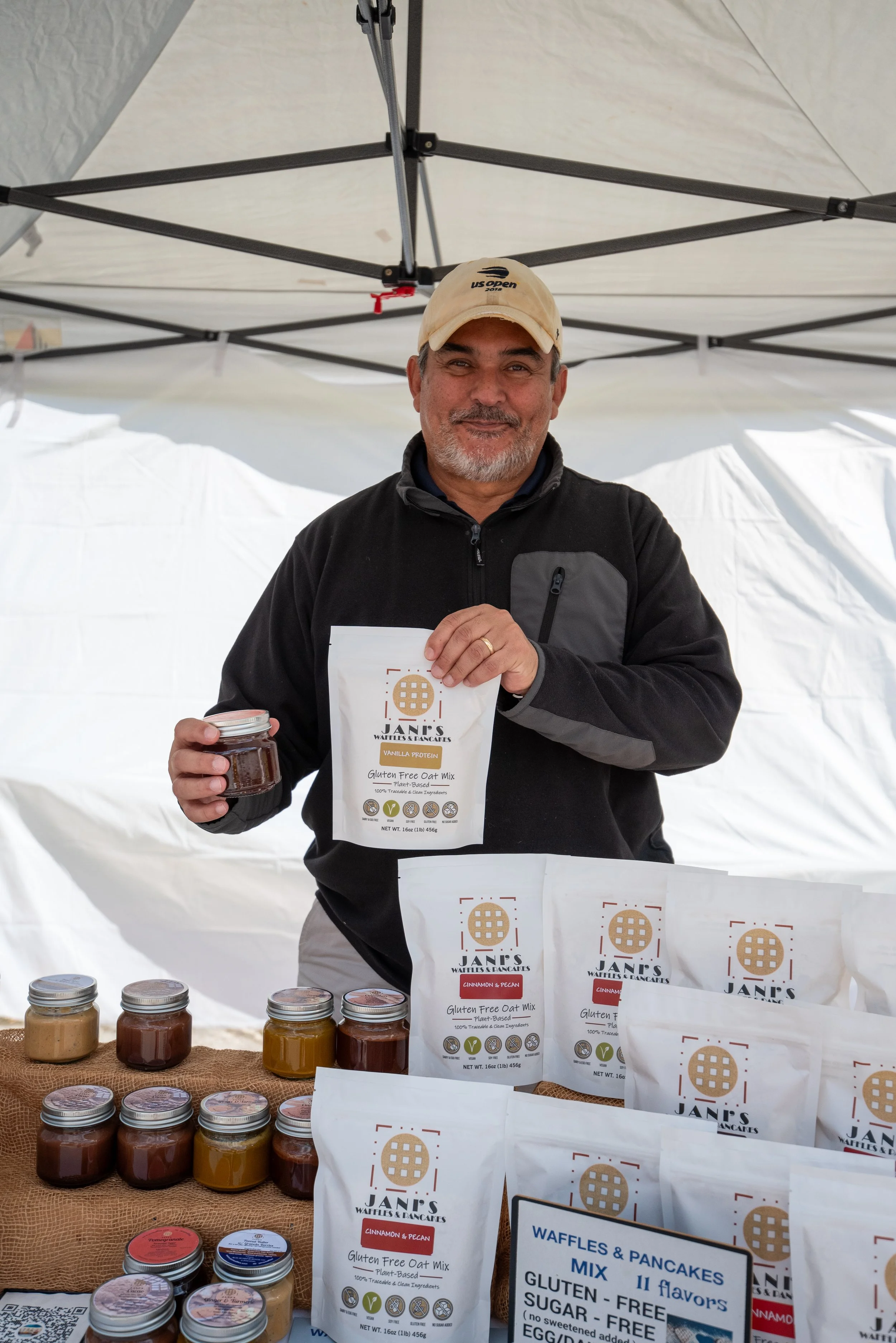 A man standing behind a table at a market stall, holding a jar and a package of gluten-free oat mix; the table displays jars of jam and packages of pancake mix, with a white tent canopy overhead.