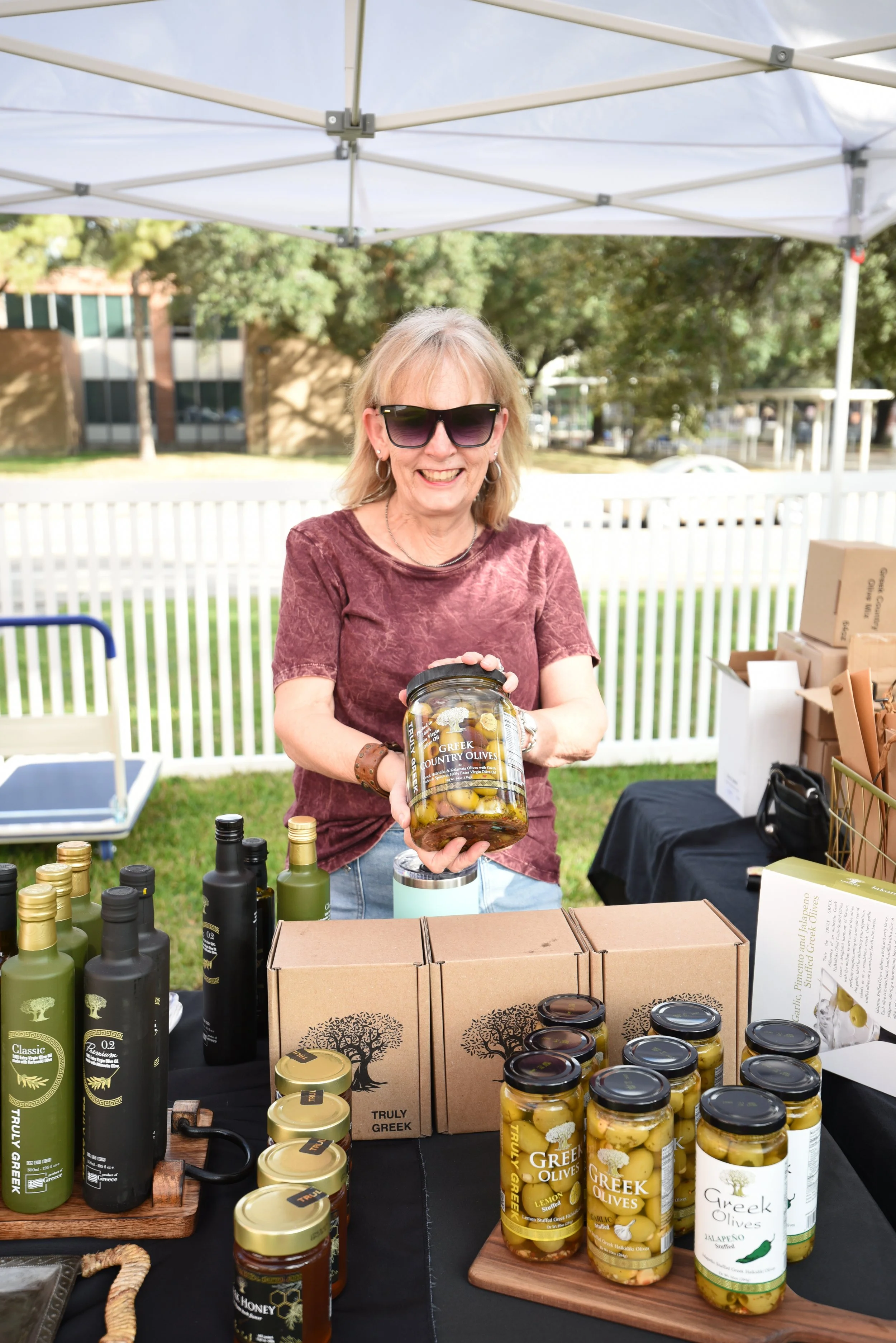 A woman with blonde hair, wearing sunglasses and a maroon shirt, holding a jar of Greek-style olives at an outdoor market stall with various bottles and jars of olives and olive products displayed on the table.