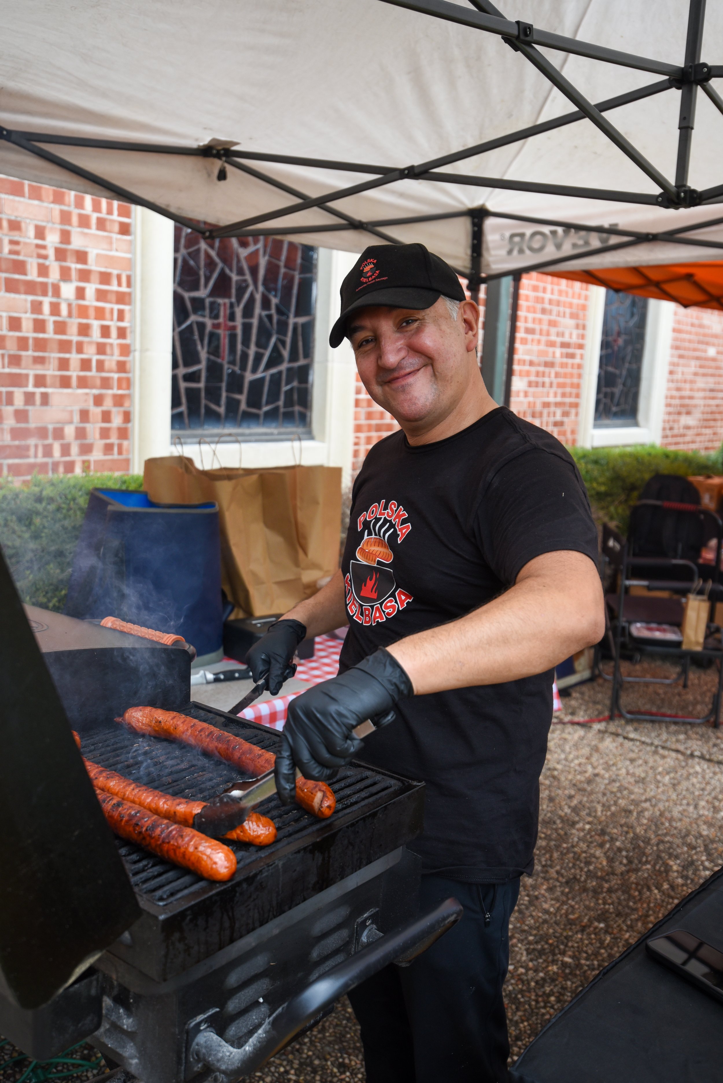 Two people smiling at a barbecue grill at an outdoor market, with sausages and vegetables cooking.
