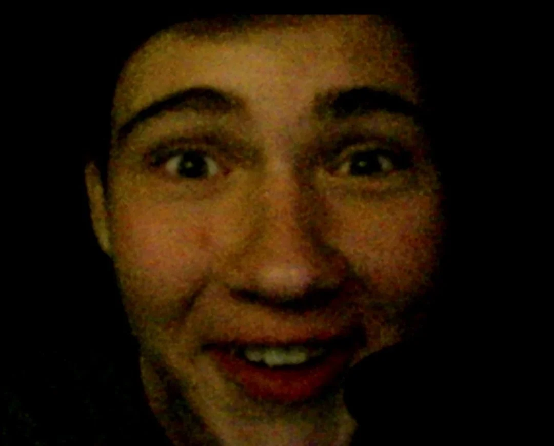 A man with dark curly hair, glasses, a beard, and a nose ring wearing a dark green sweater. He is looking slightly to his left with a neutral expression, in an indoor setting with a plant in the background.