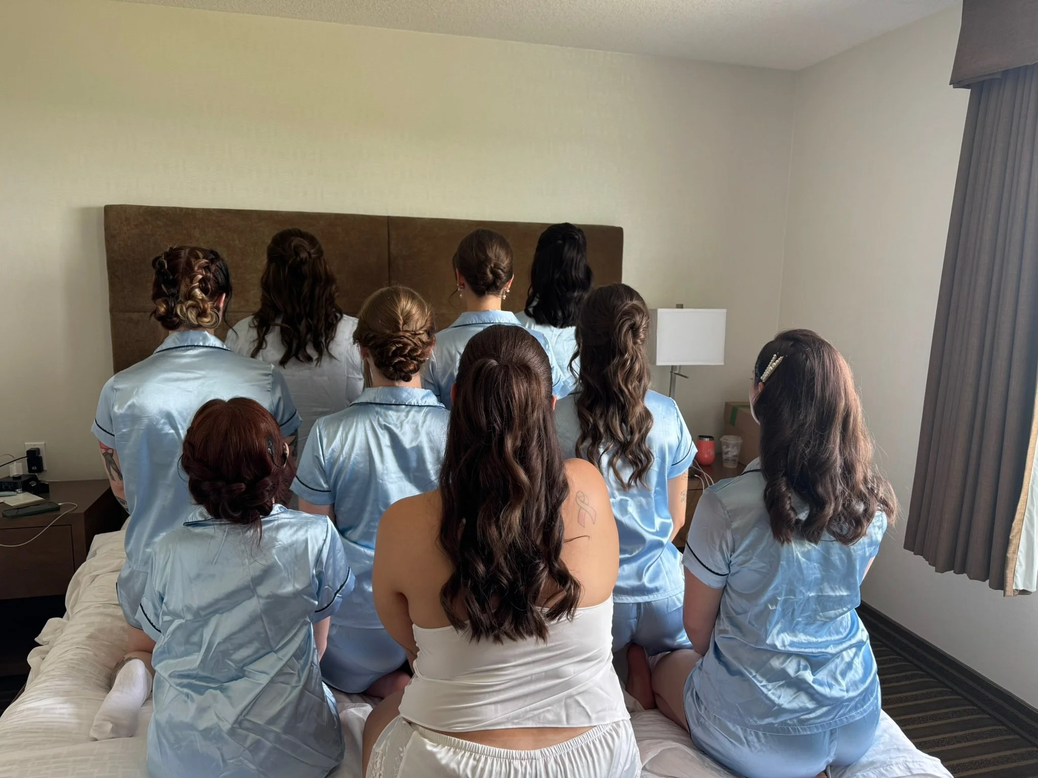 Ten women dressed in matching light blue pajamas sitting on a bed in a hotel room, facing the wall, with a woman in a white dress sitting among them.