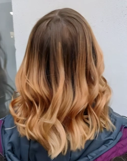 Back view of a person with shoulder-length, wavy blonde hair, wearing a dark jacket with purple accents, standing against a white wall.