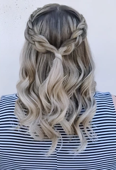 Back view of a woman with long, wavy blonde hair styled in a half-up, twisted crown braid, wearing a striped shirt.