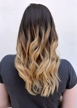 Back view of a woman with long, wavy hair that transitions from dark brown at the roots to blonde at the tips, wearing a dark grey top.
