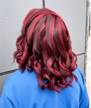 Back view of a woman with shoulder-length, curly hair dyed in shades of red and pink, wearing a blue top.