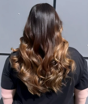 Back view of a woman with long, wavy brown hair with blonde highlights, wearing a black shirt.