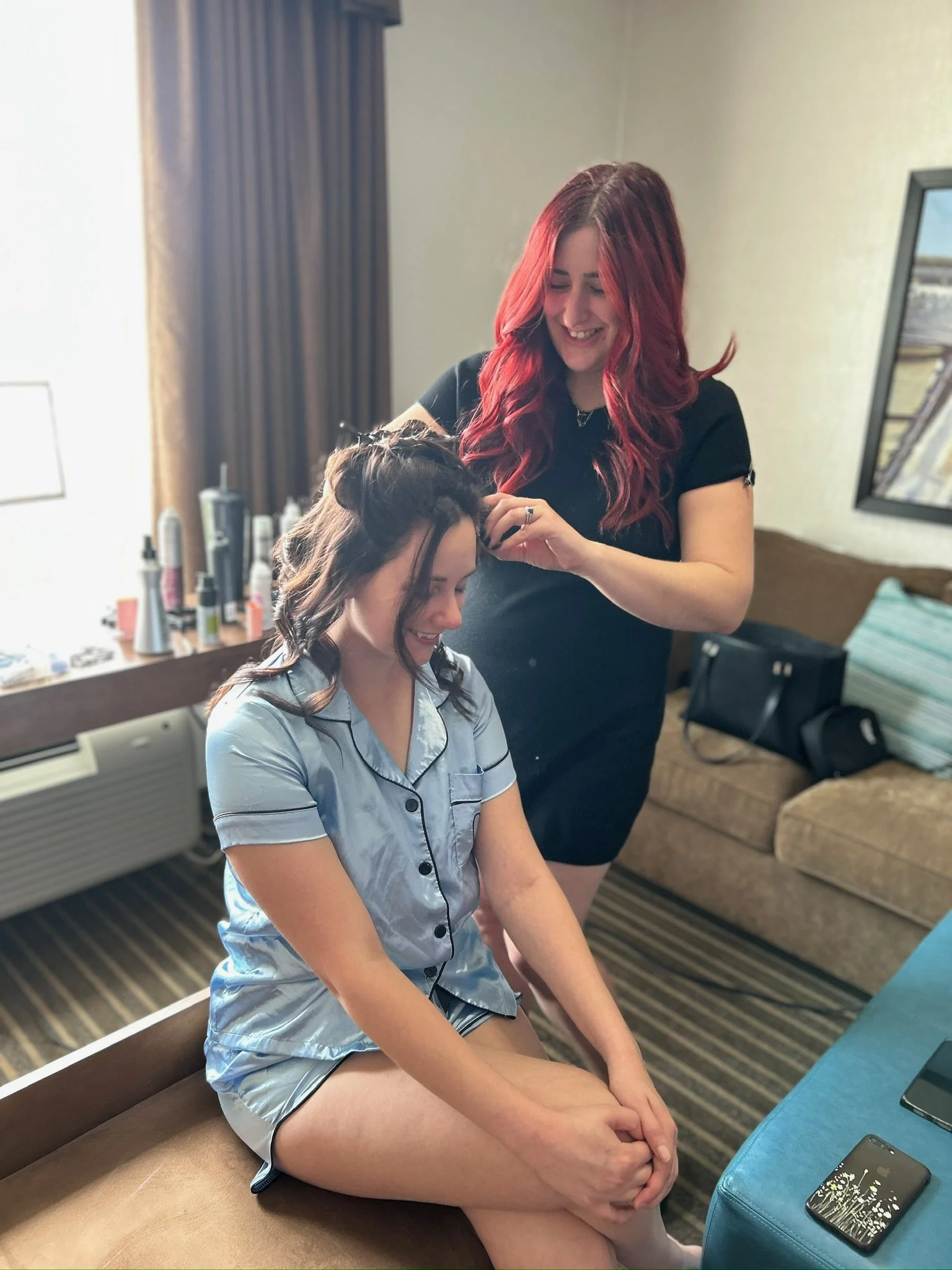 A woman sitting on a bed with short pajamas, having her hair styled by another woman in a hotel room.