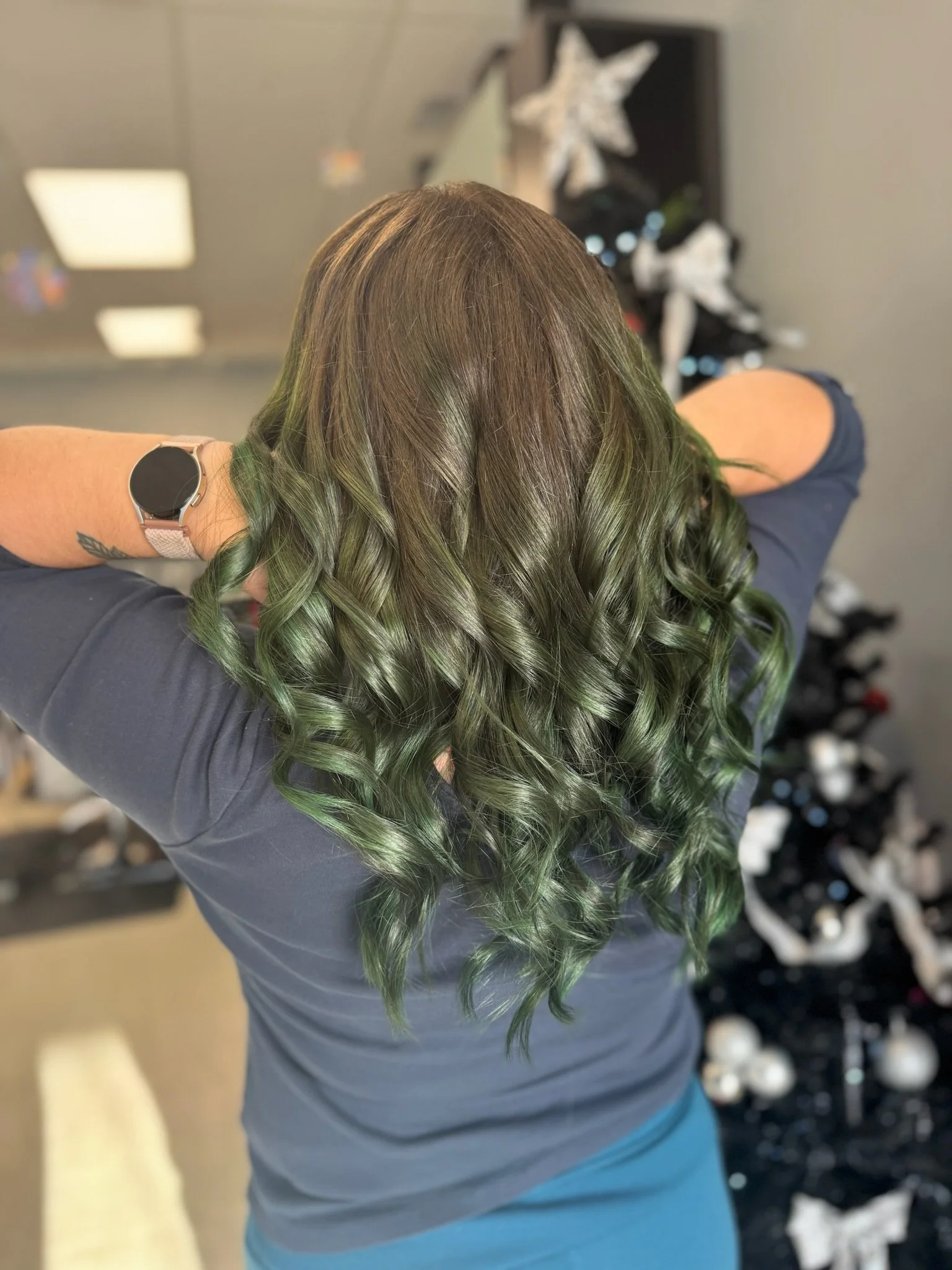 Back view of a woman with green and brown curly hair, wearing a dark gray top and a fitness tracker on her wrist, in a salon or similar setting.