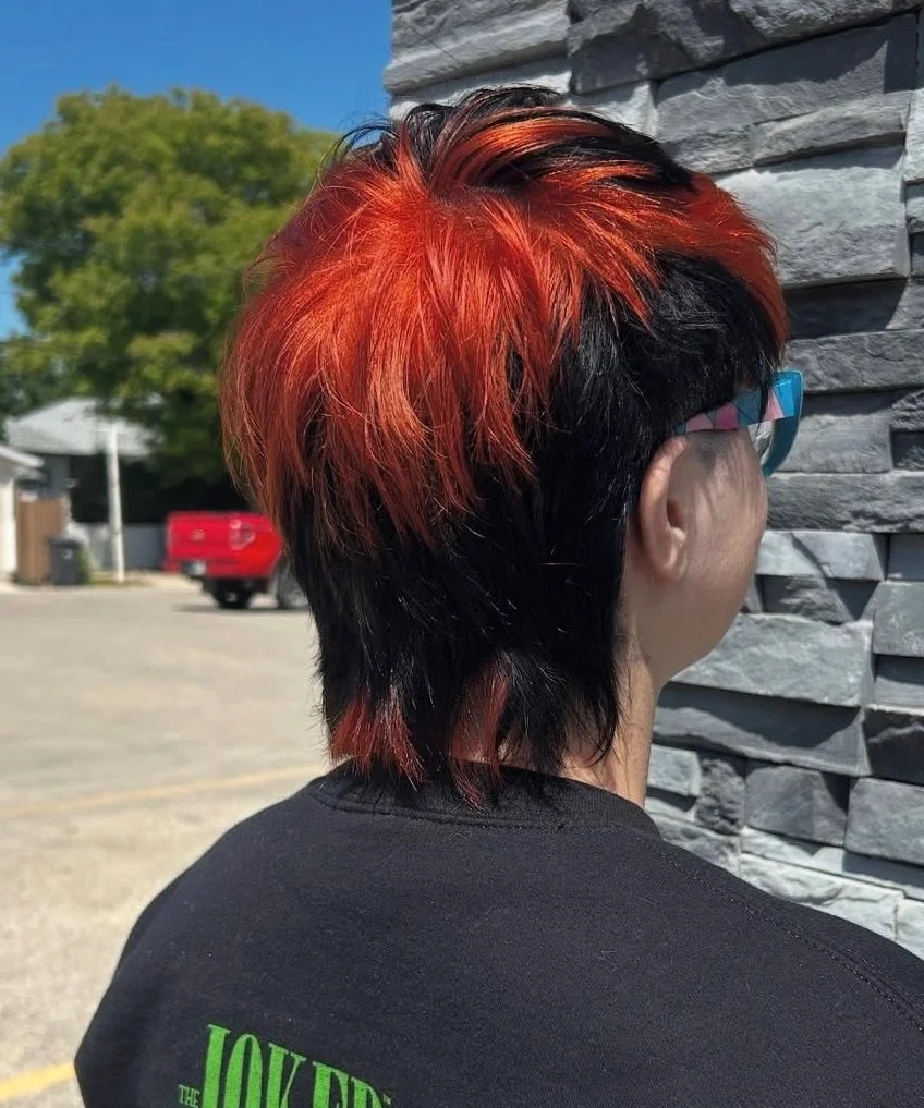 Person with short layered hairstyle featuring vibrant red and black hair, wearing colorful glasses, standing outdoors against a stone wall.