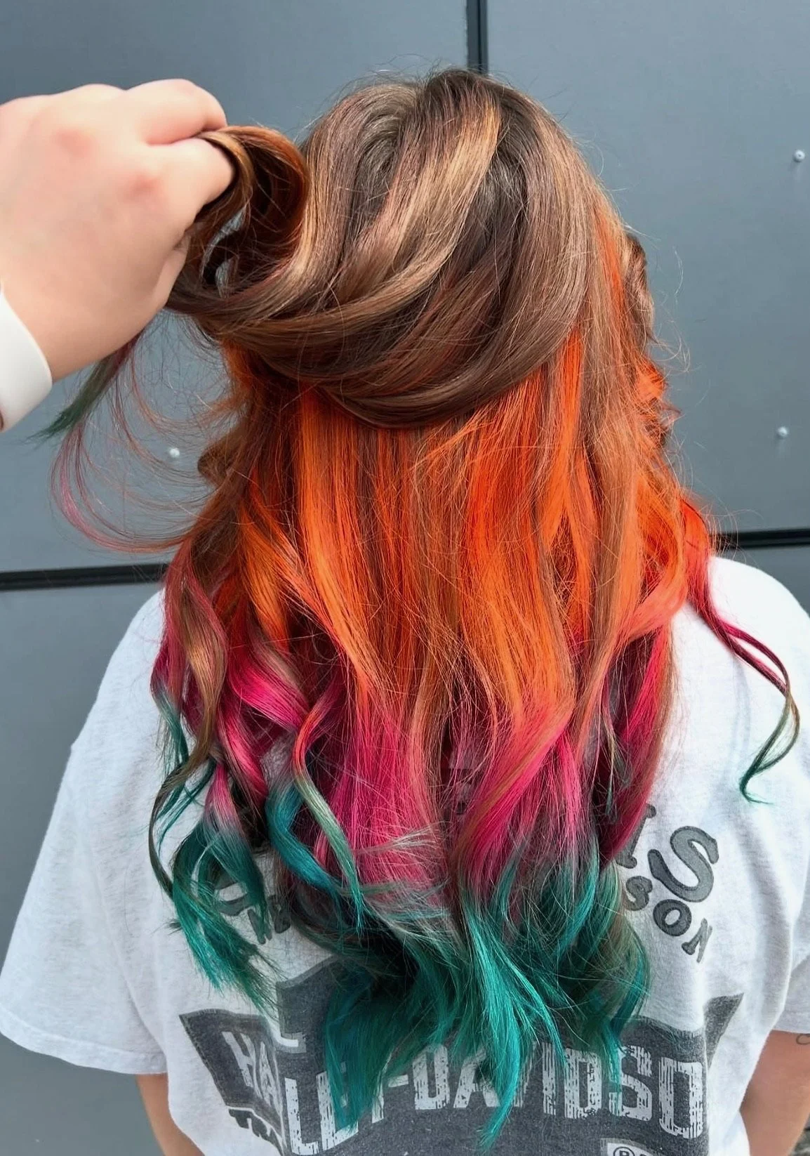 A woman with long, wavy hair dyed in multiple vibrant colors, including orange, pink, green, and blue, standing against a gray background.