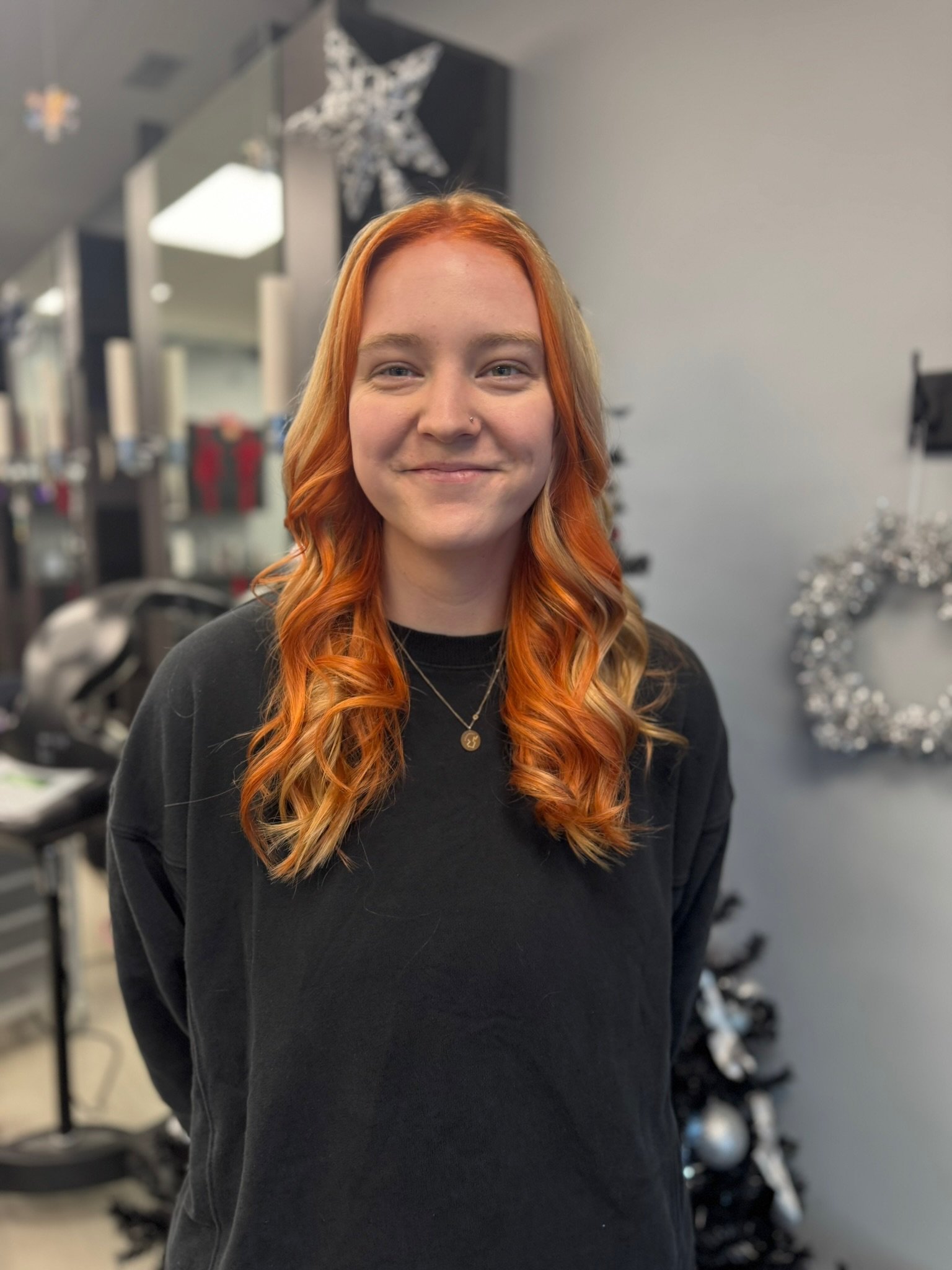 A young woman with red hair styled in loose curls, wearing a black sweatshirt and small necklace, standing indoors with holiday decorations in the background.