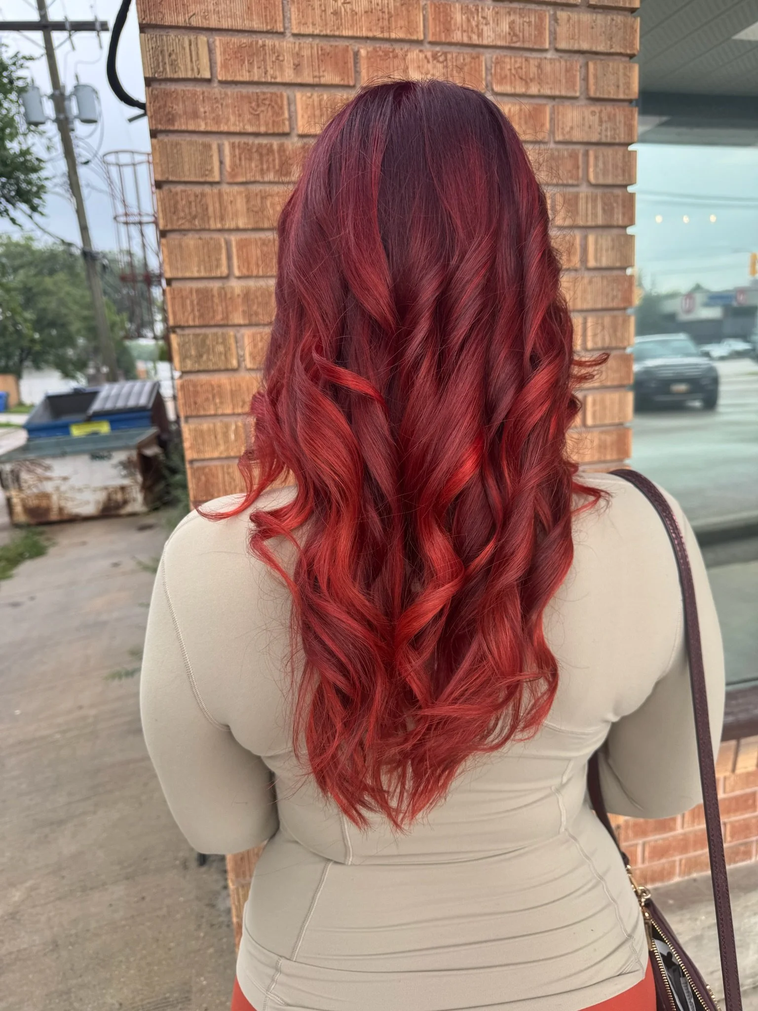 Back view of a woman with long, curly, vibrant red hair standing outside near a brick wall, with a parking lot and a few buildings in the background.