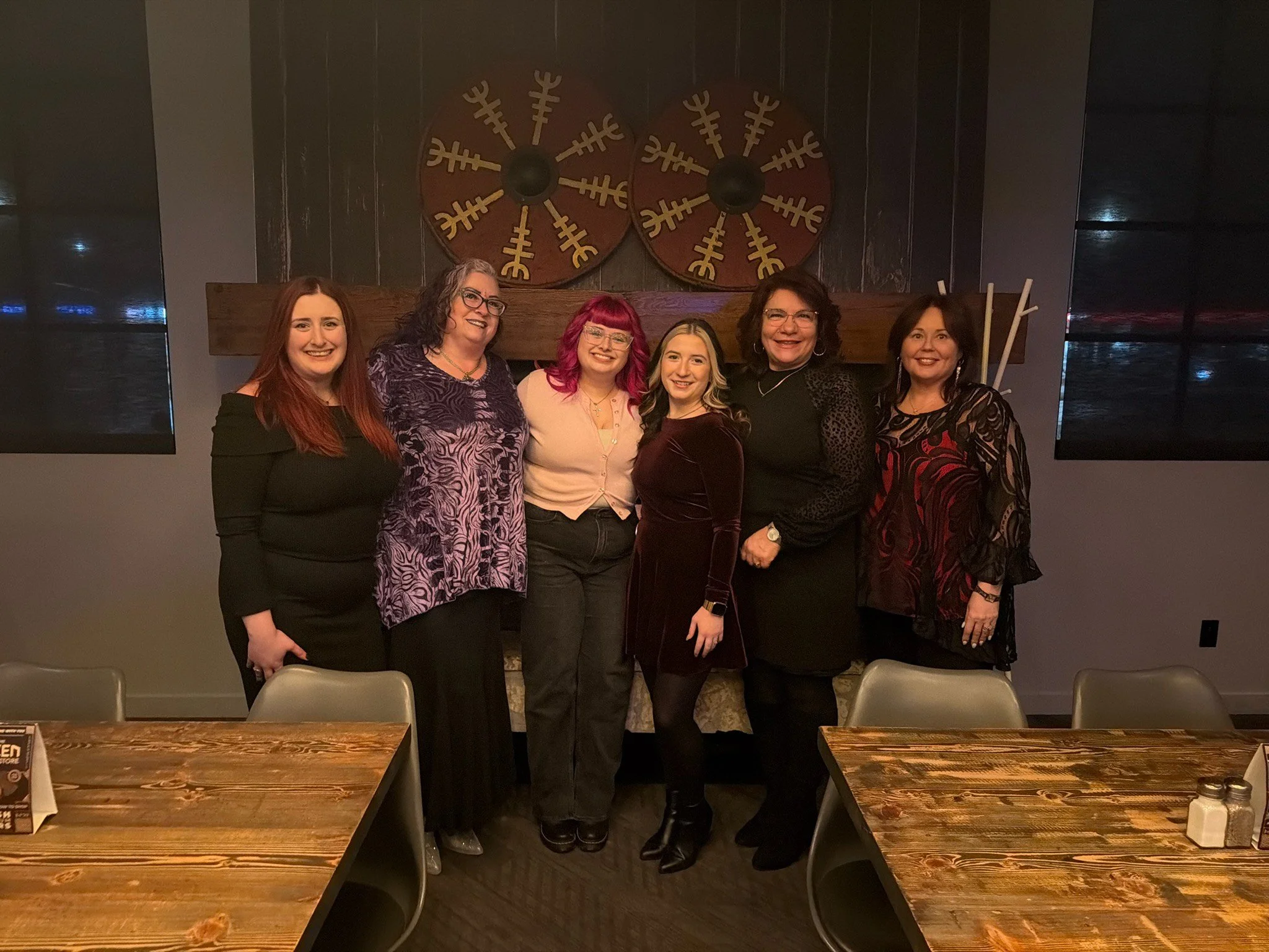 Six women standing together in a restaurant or event space, smiling at the camera, with a wooden fireplace mantel and decorative wall art behind them.