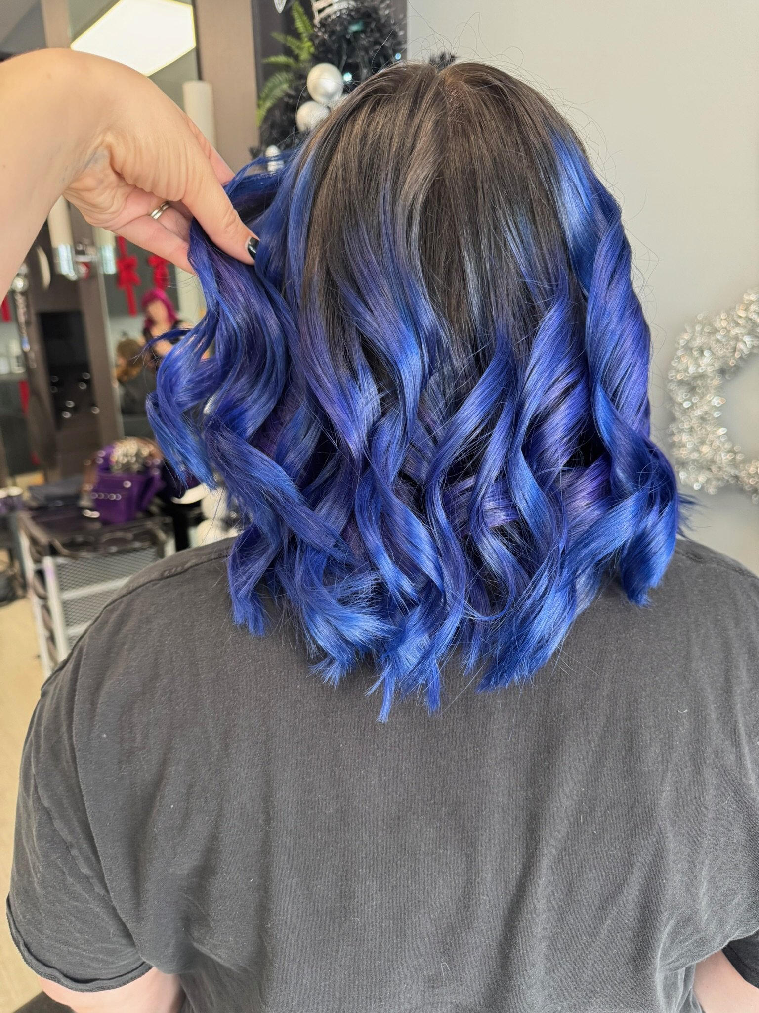 Back view of a person with shoulder-length dark hair with blue highlights, styled in loose curls, in a hair salon.