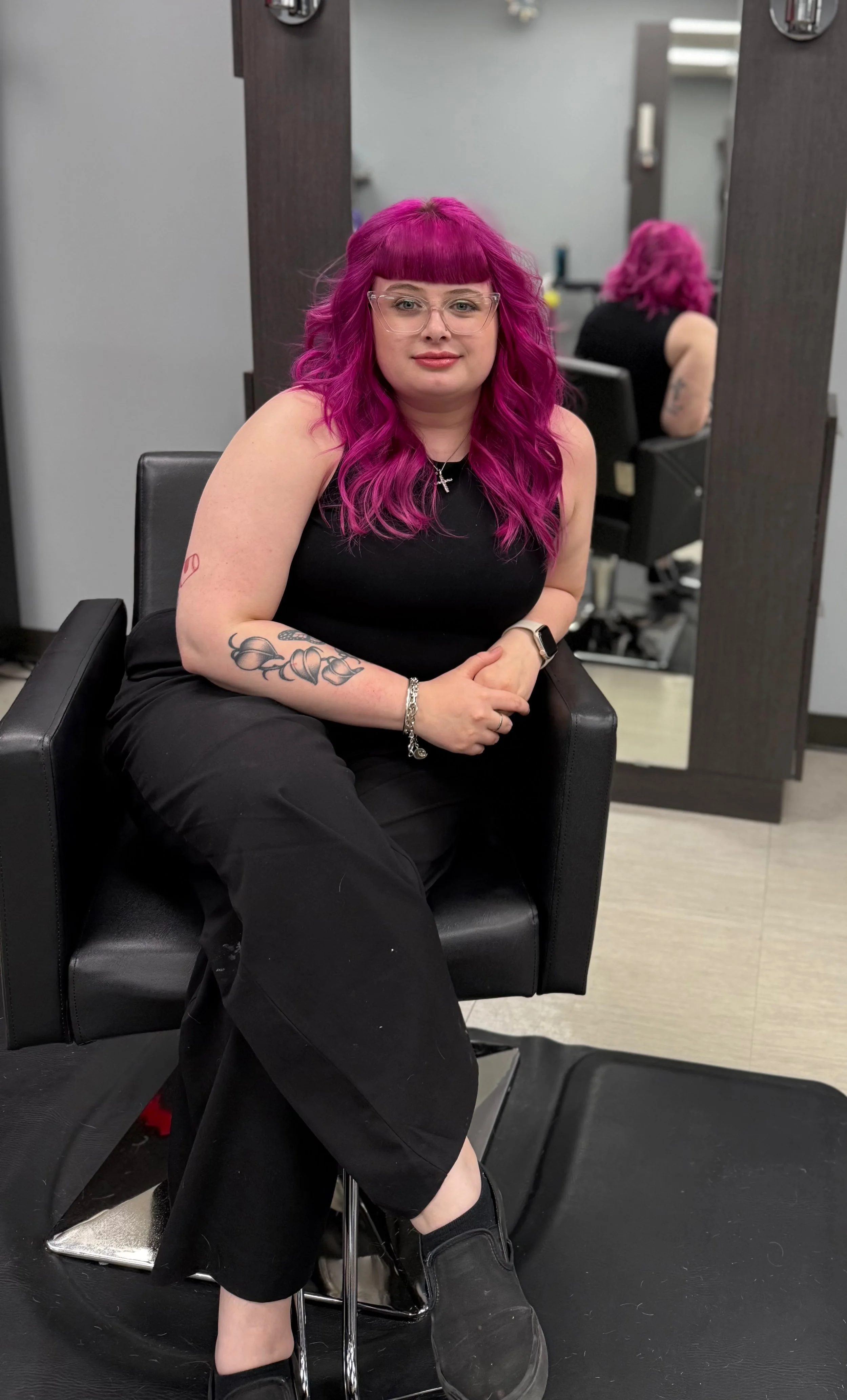 A woman with bright pink hair and glasses sitting in a black salon chair at a hair salon.