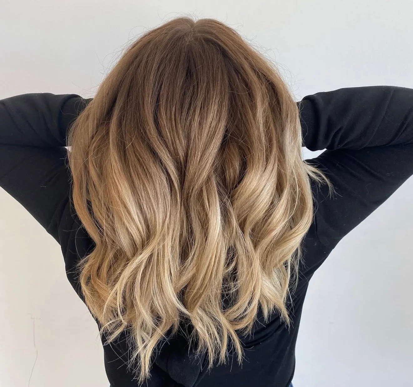 Person with long, wavy, blonde hair, wearing a black top, with arms raised behind head, against a plain white background.
