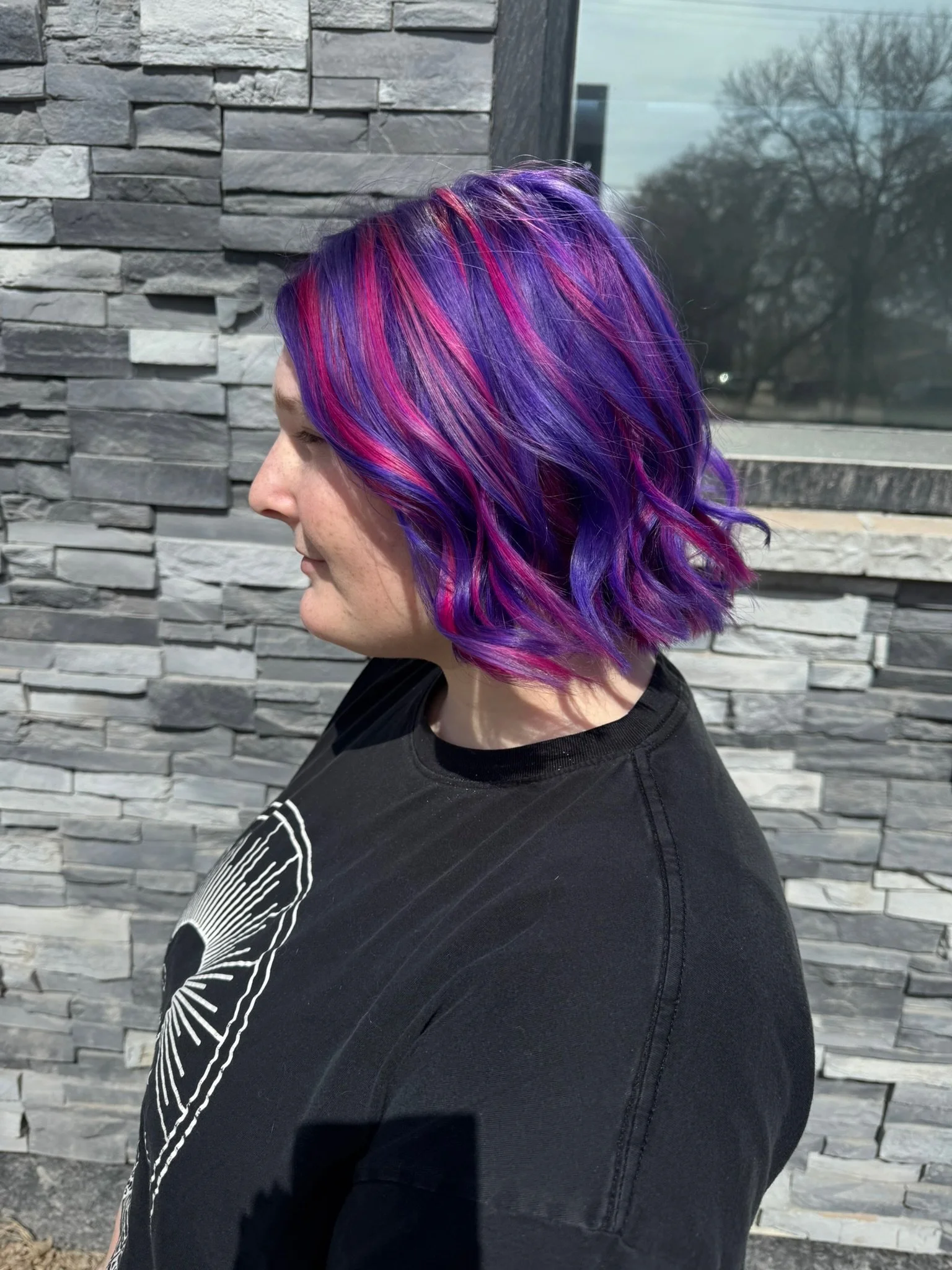 A woman with chin-length wavy hair dyed in shades of purple and pink, standing against a stone wall exterior with a window and trees in the background.