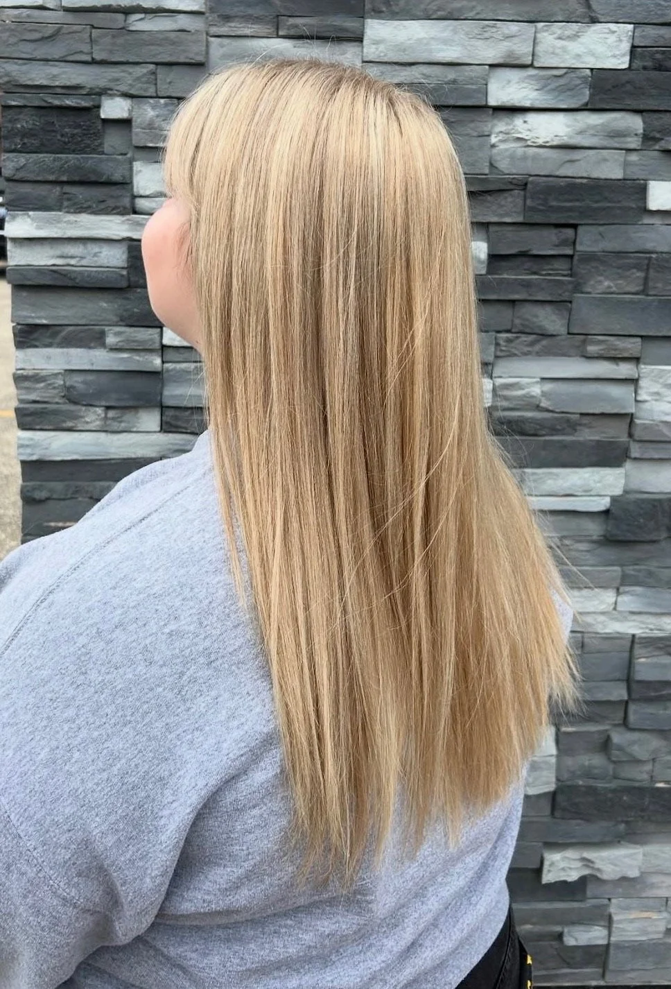 Back view of a woman with long, straight blonde hair, wearing a gray shirt, standing in front of a textured gray stone wall.