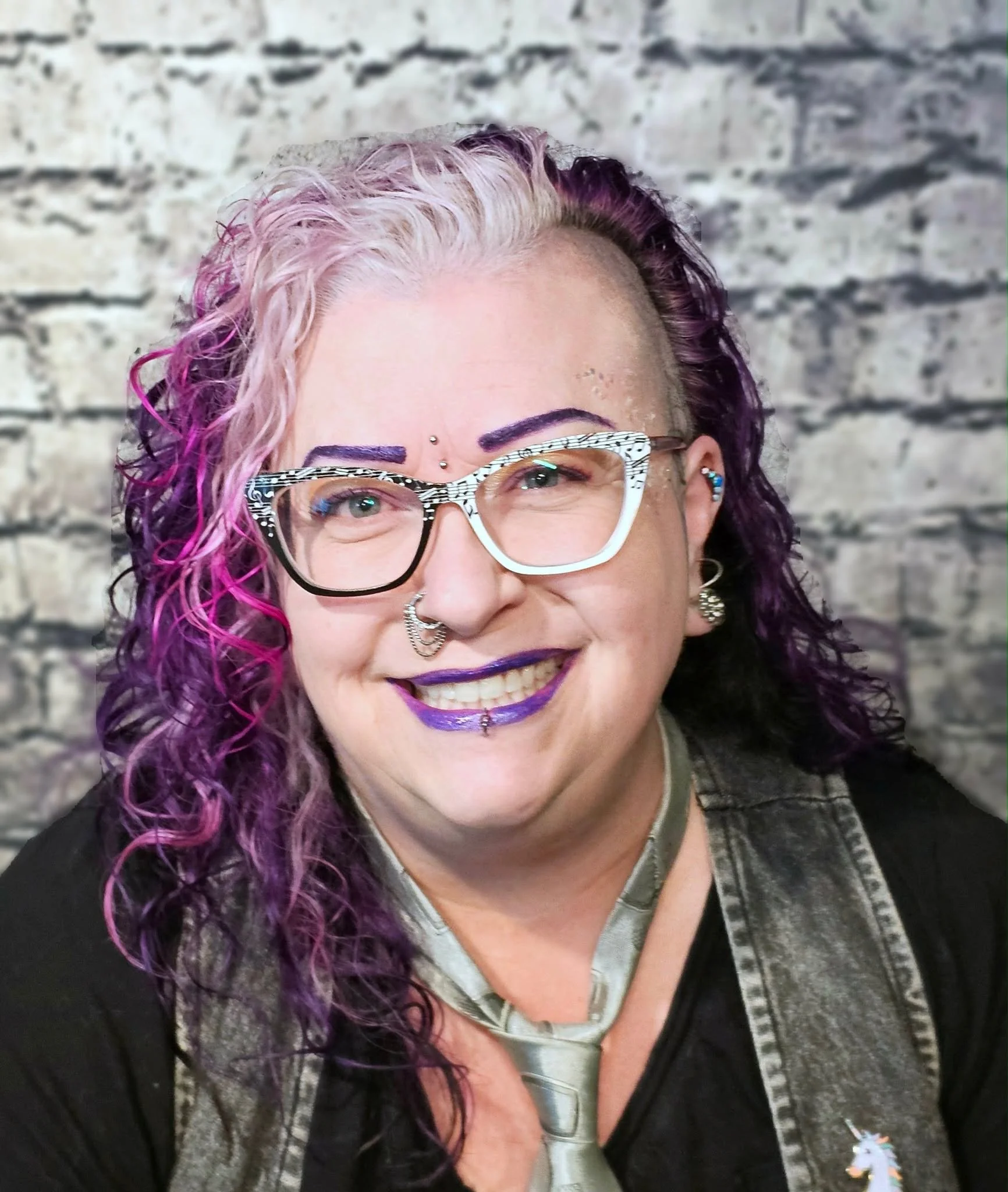 A woman with pink and purple curly hair, wearing large glasses with a white and black pattern, purple lipstick, nose and lip piercings, earrings, and a unicorn pin on her vest, standing against a textured brick wall.