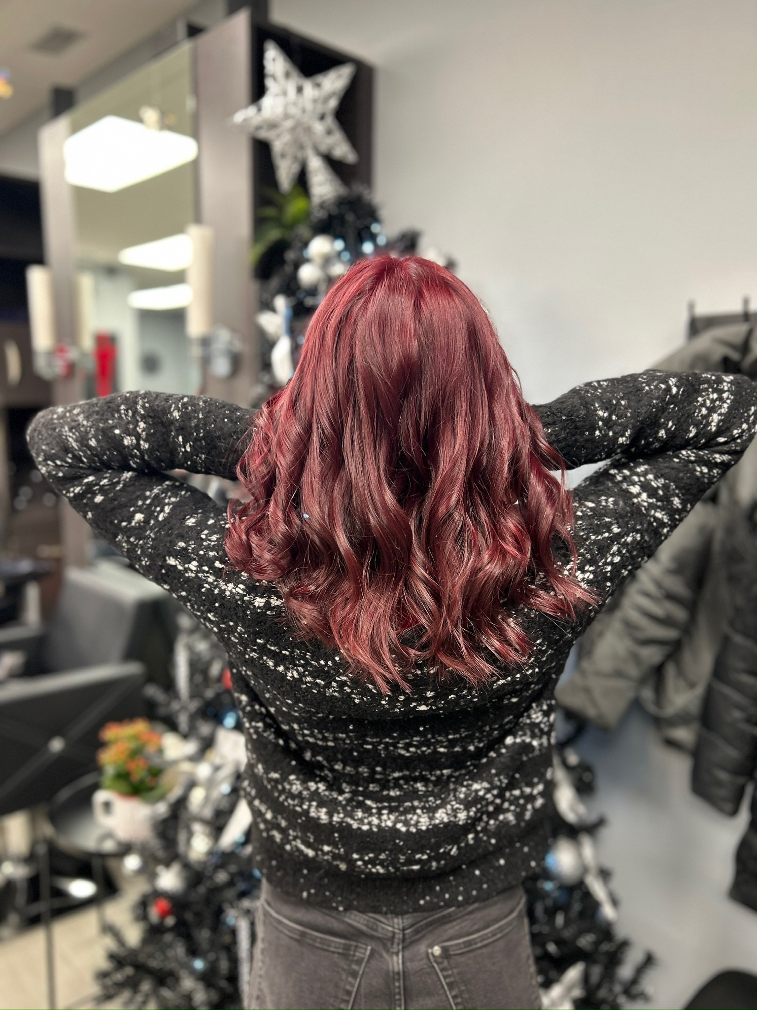 A woman with wavy, red hair, wearing a black sweater with white speckles, poses with her back to the camera in a room decorated for Christmas.