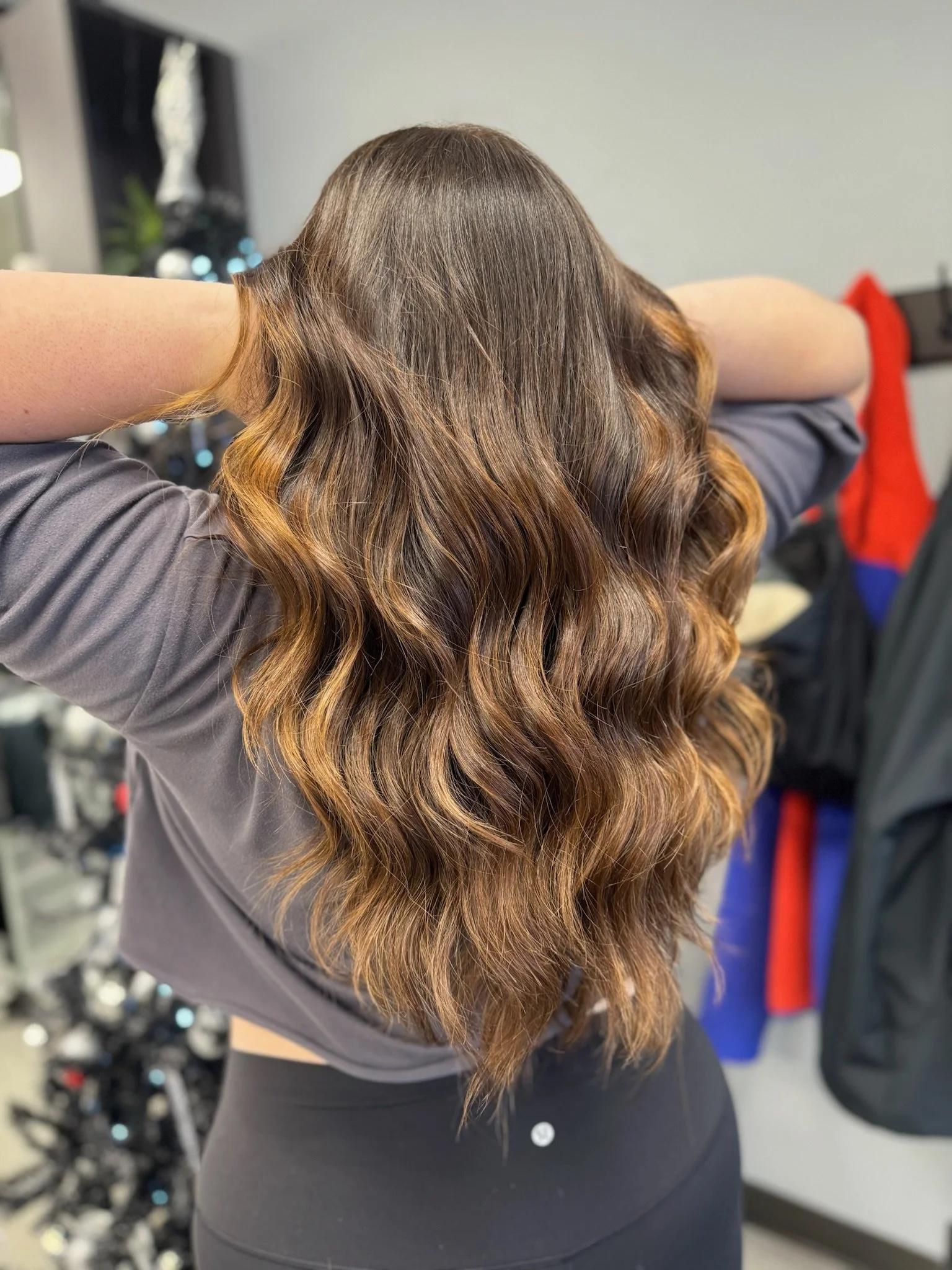 Back view of a woman with long, wavy brown hair with caramel highlights, wearing a gray top, in a salon or studio setting.