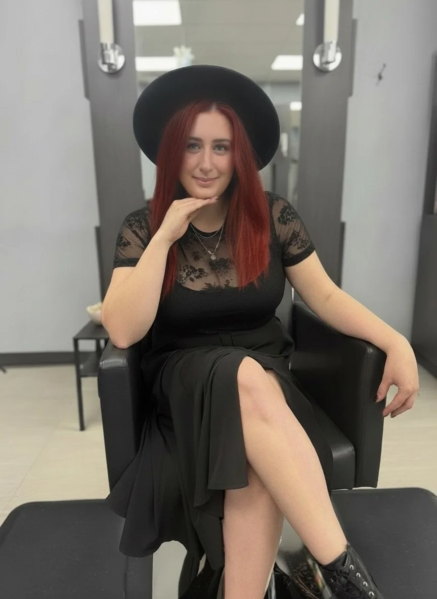 A woman with red hair, wearing a black hat and a black lace top, sitting in a salon chair with one leg crossed over the other, inside a salon.