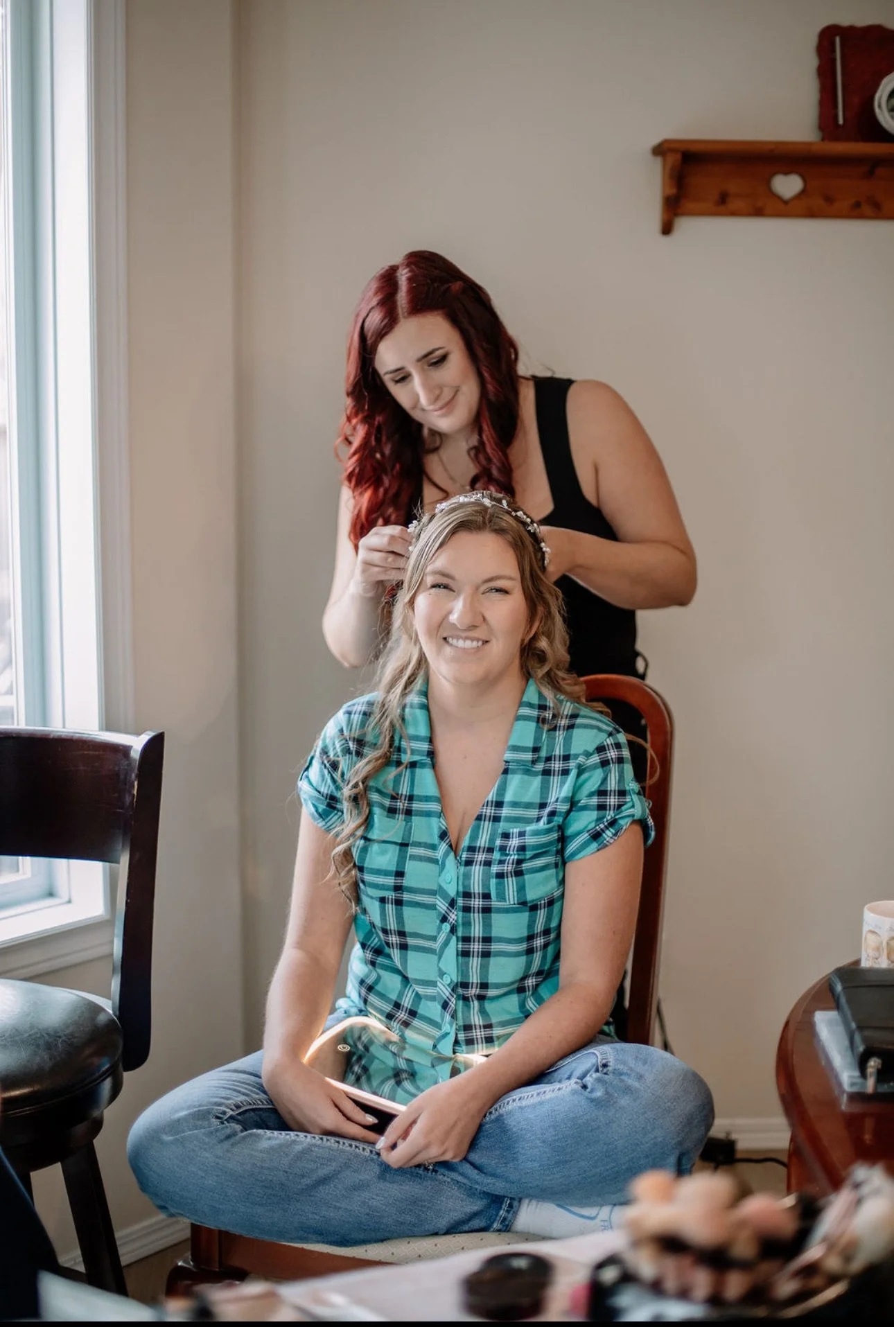 A woman in a blue plaid shirt sitting cross-legged and smiling while having her hair done by another woman with red hair, in a well-lit room.