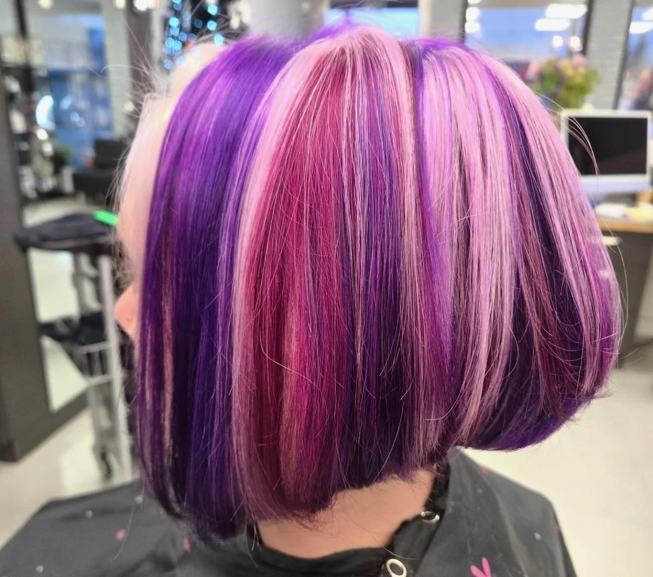 Back view of a person's short, straight hair dyed in vibrant shades of purple, pink, and blonde with distinct streaks, in a salon setting.