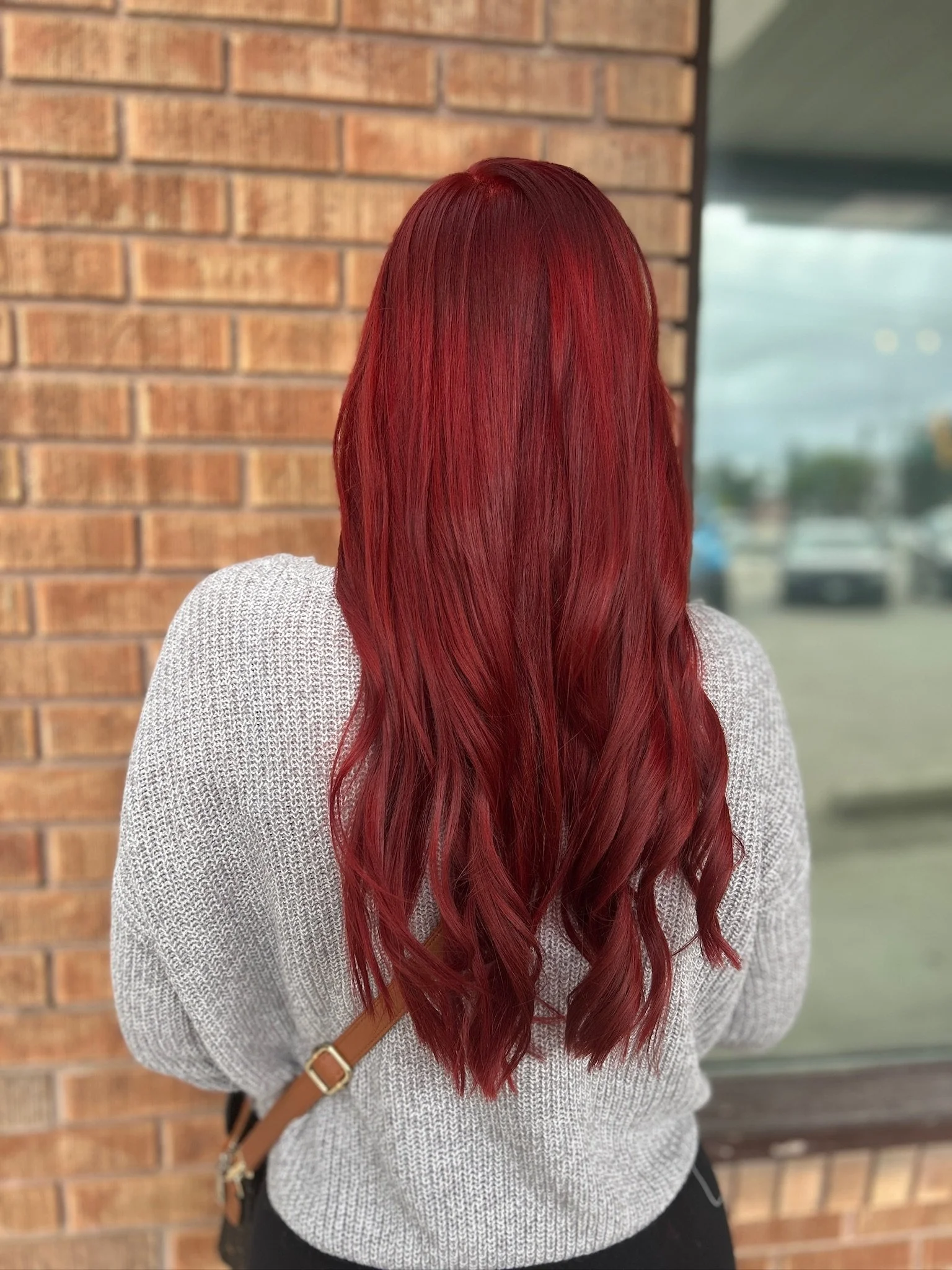 Back view of a woman with long, wavy, vibrant red hair, standing outside near a brick wall and a window.