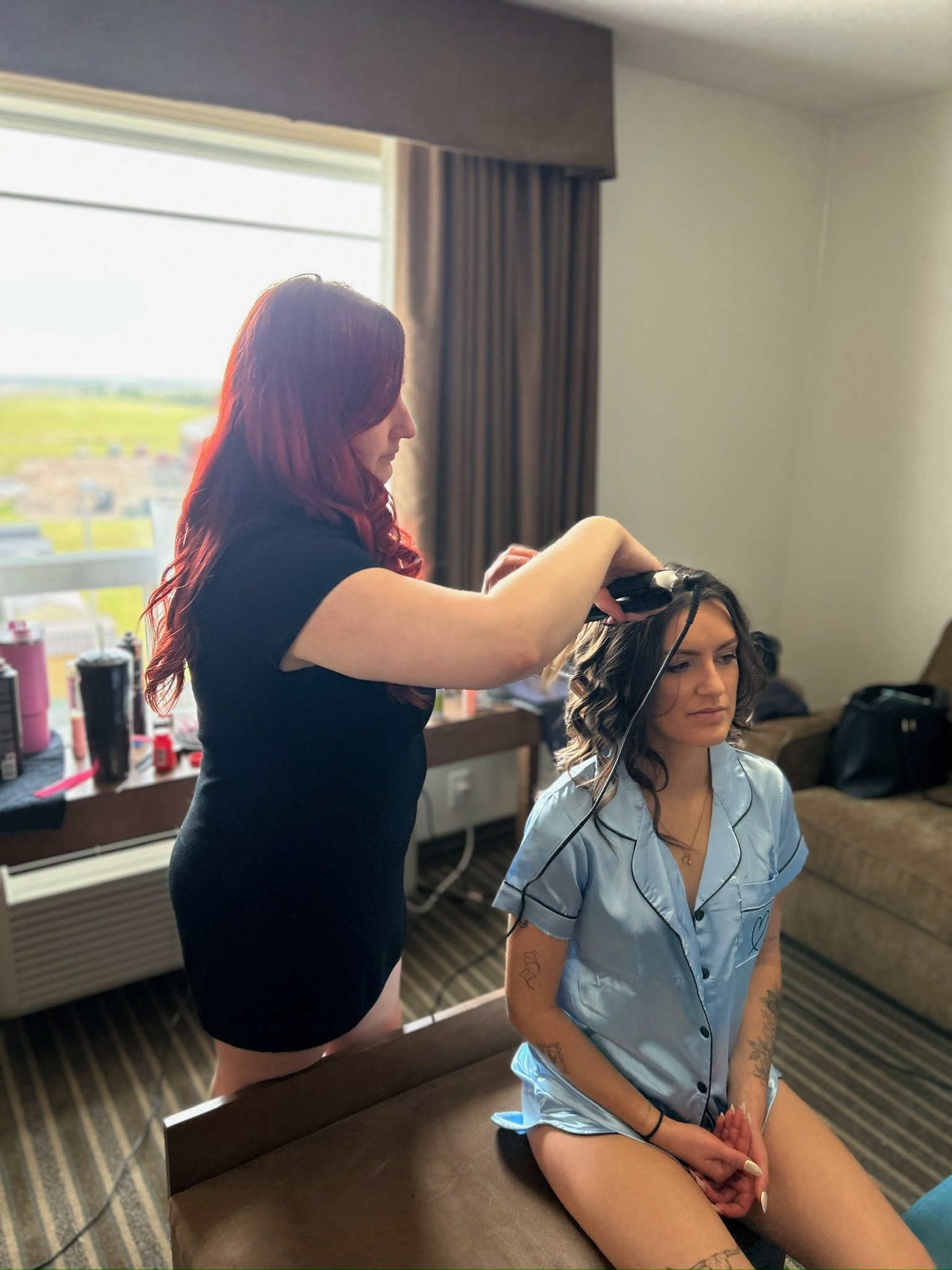 Woman with red hair styles a woman with dark hair sitting on a bed in a hotel room.