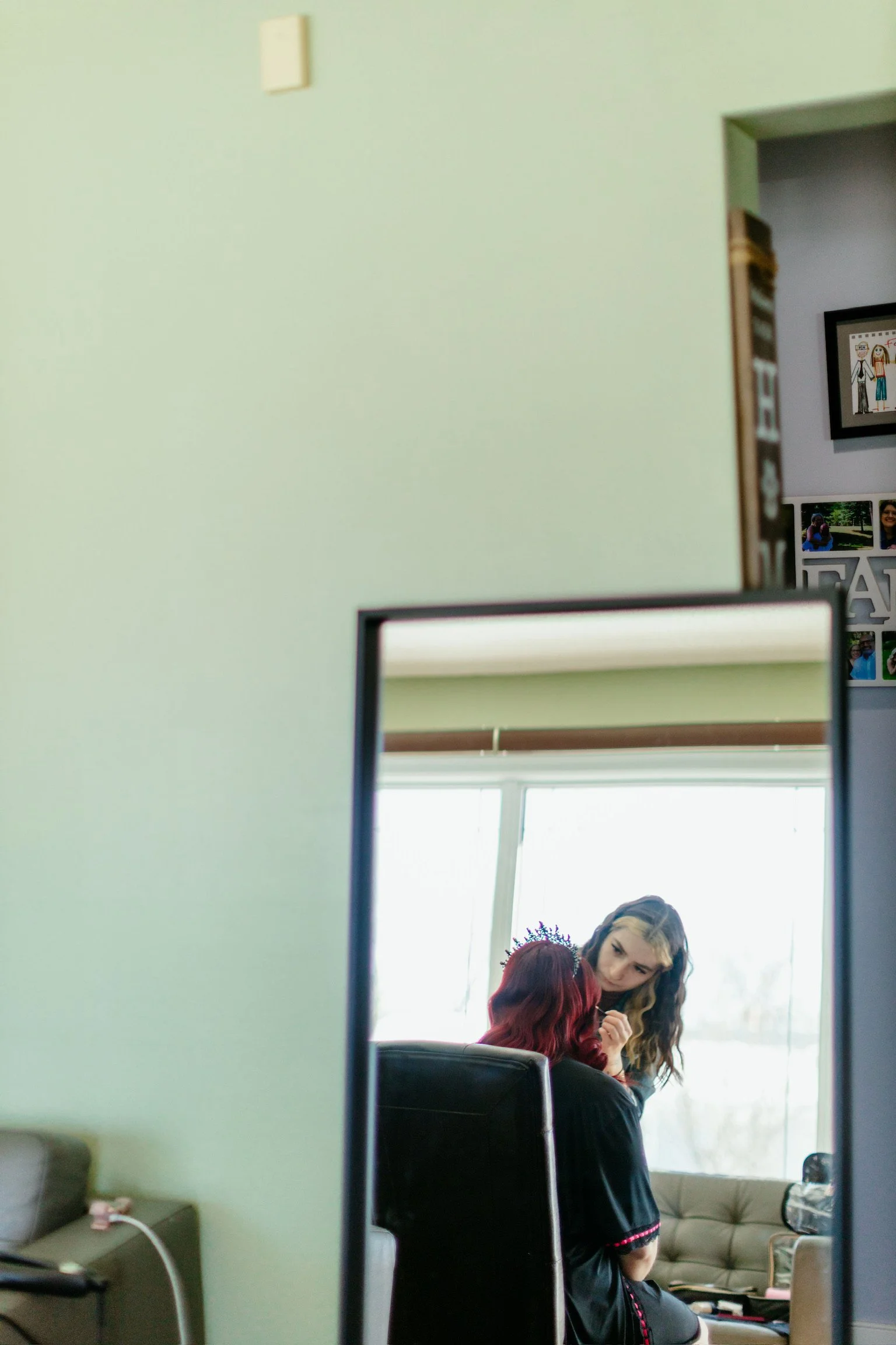 A mirror reflecting a woman with long hair and a black dress with pink trim, leaning in towards a young girl with red hair and a purple headband, in a room with a large window and a beige couch.
