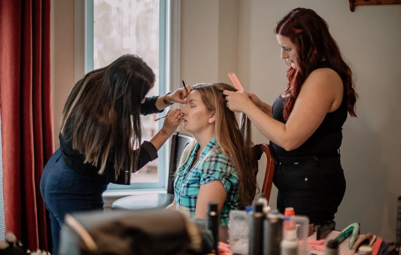 A woman sitting with her eyes closed as two makeup artists apply makeup and style her hair in a room with natural light.