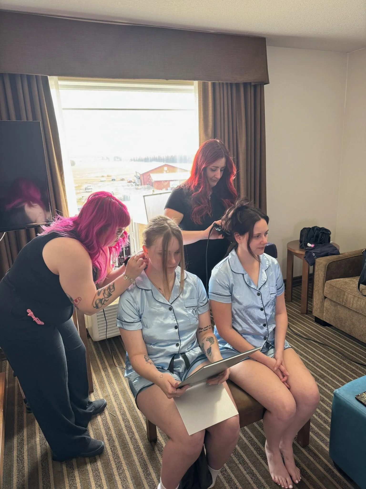 Two women sitting on a chair reading a scrapbook while two hairstylists do their hair in a hotel room, with a large window and a TV in the background.