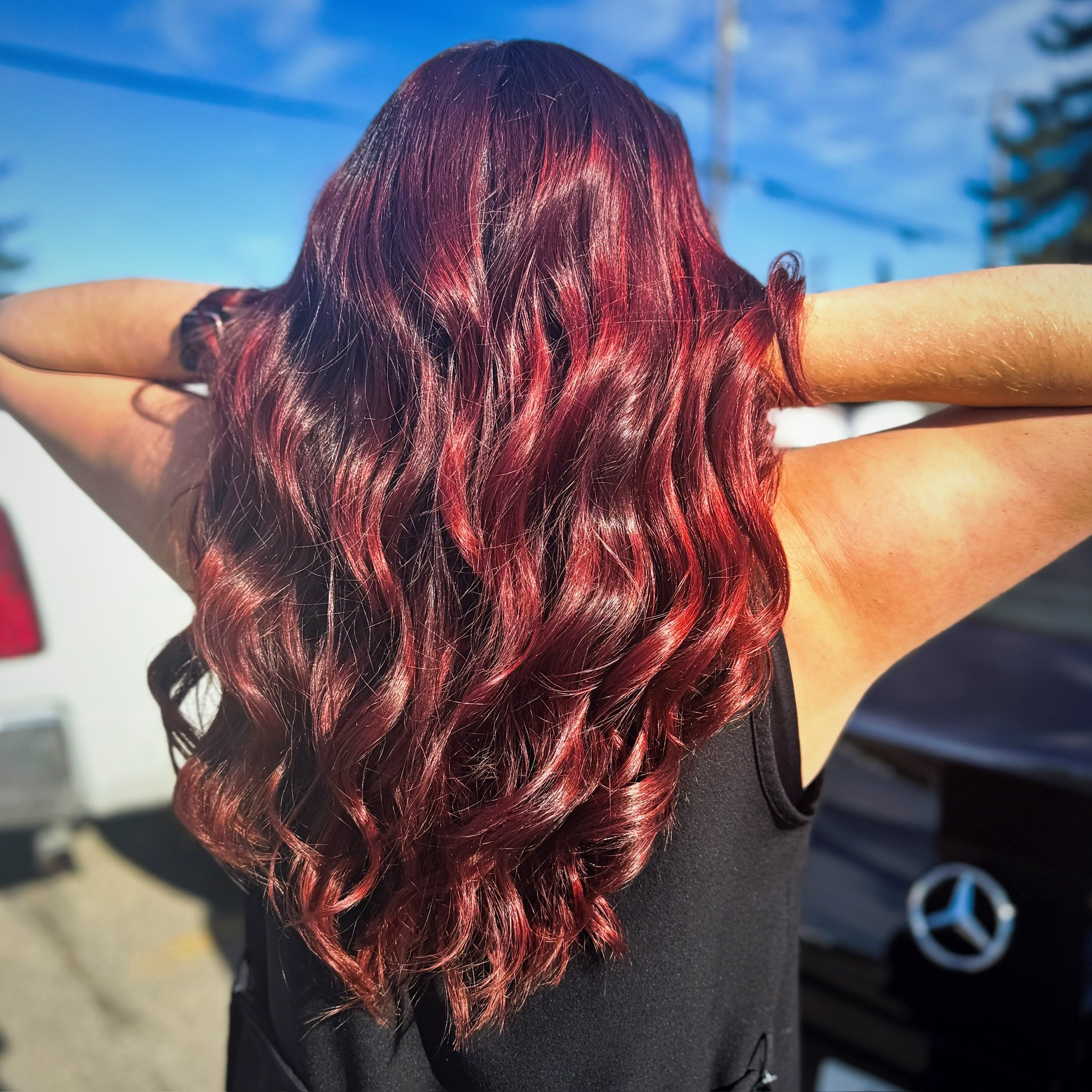 Back view of a woman with long, wavy, red hair, wearing a black sleeveless top, outdoors on a sunny day.