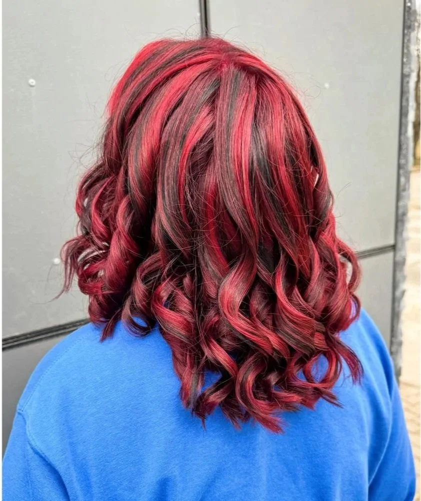 Back view of a person with wavy, shoulder-length hair dyed in shades of red and black, wearing a blue top.