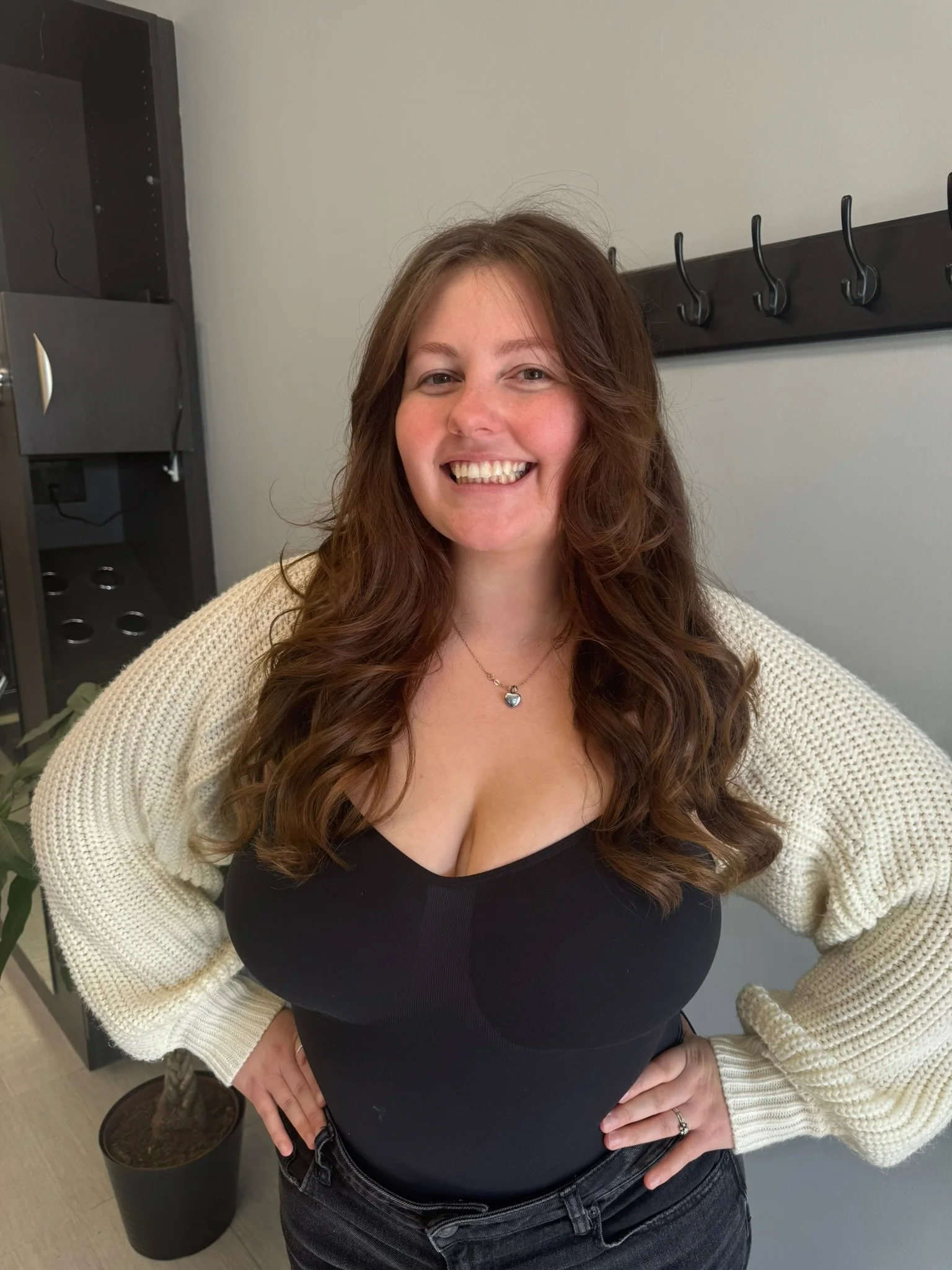 Smiling woman with long, wavy brown hair, wearing a black top and cream-colored cardigan, standing indoors with hands on hips.