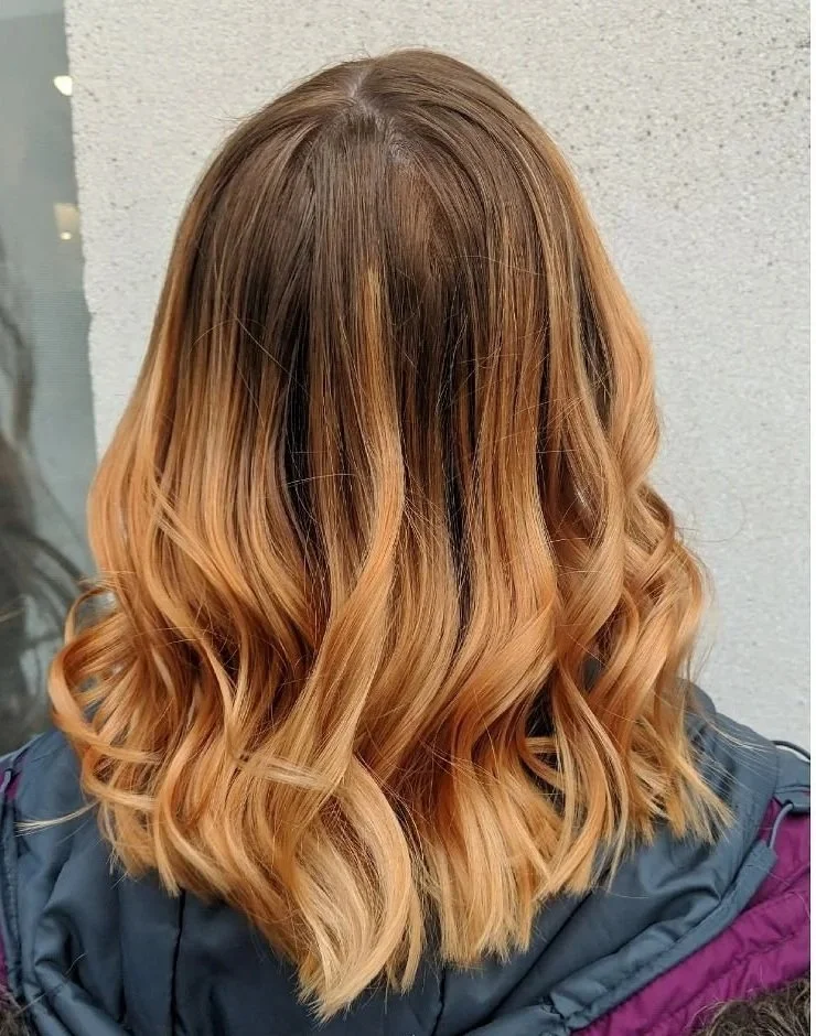 Back view of a woman with shoulder-length, wavy hair that is balayaged with darker roots fading into warm, honey blonde ends.