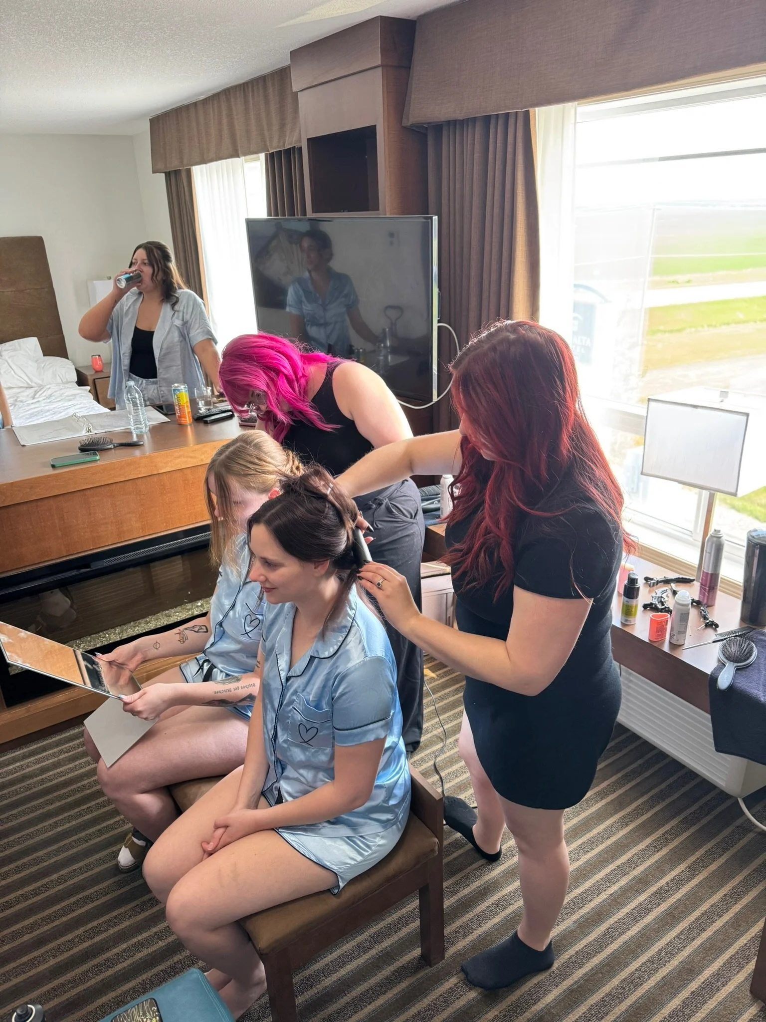 A woman with red hair styles a woman with dark hair and tattoos at a hotel room vanity while a woman with pink hair styles her hair, with others in pajamas, some sitting and some standing.