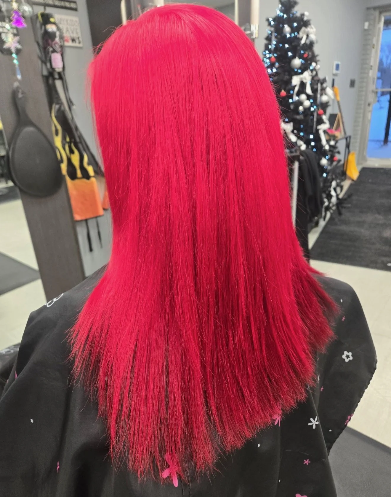 Back of a person's head with bright red, straight, shoulder-length hair in a hair salon. In the background, there is a decorated Christmas tree and various salon tools.