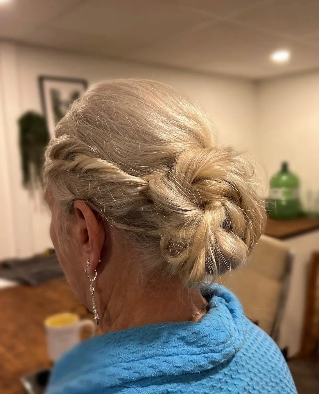 Back view of an elderly woman with blonde hair styled in an elegant updo, wearing a blue garment and earrings, in a home setting.