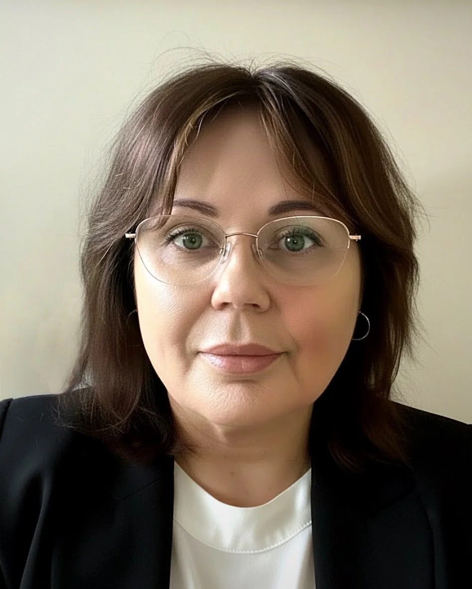 A woman with shoulder-length brown hair, wearing glasses, earrings, a black blazer, and a white top, looking directly at the camera against a plain background.