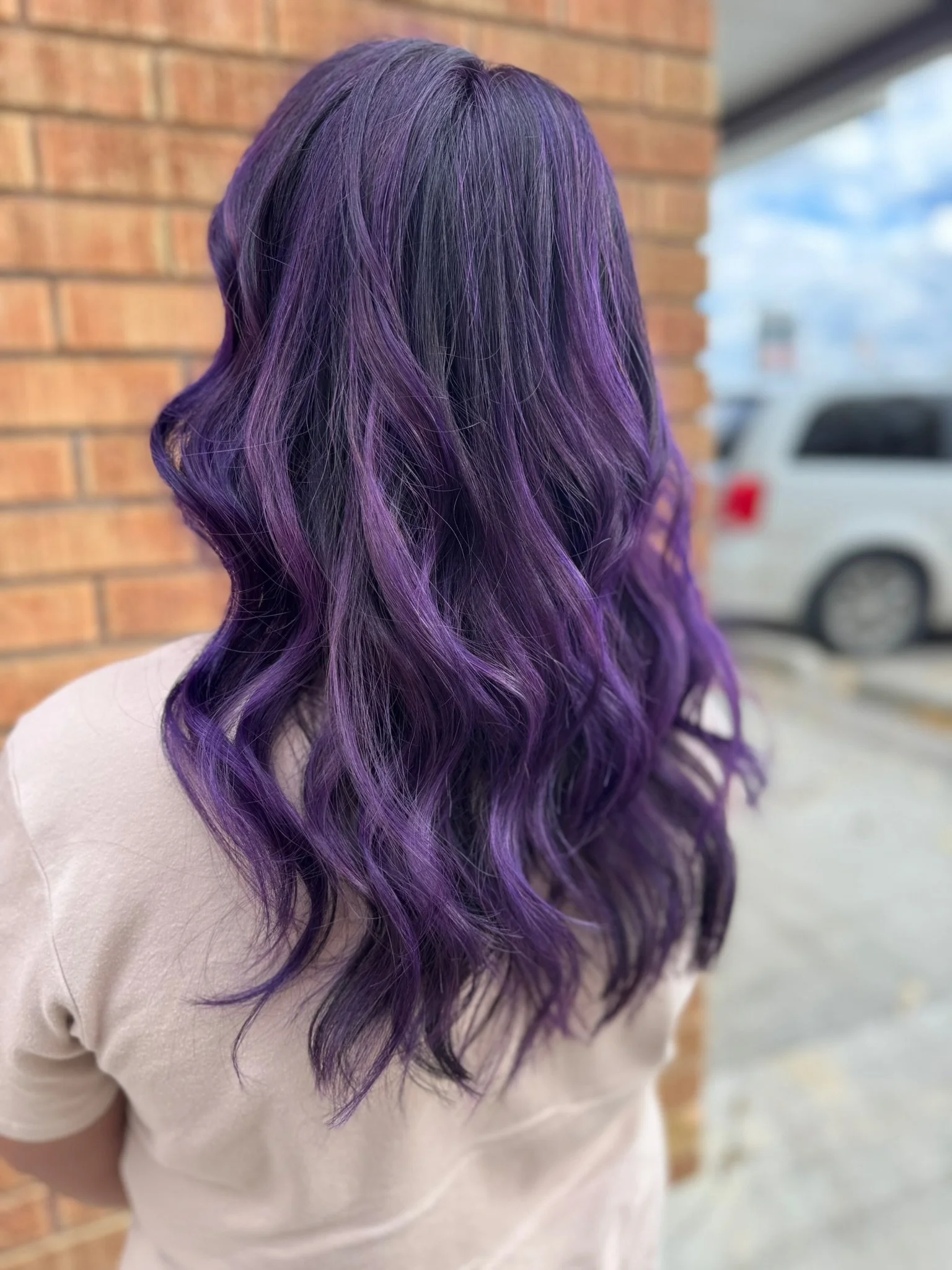 Woman with wavy purple and black ombre hair standing outdoors near a brick wall, with a cloudy sky and a vehicle in the background.
