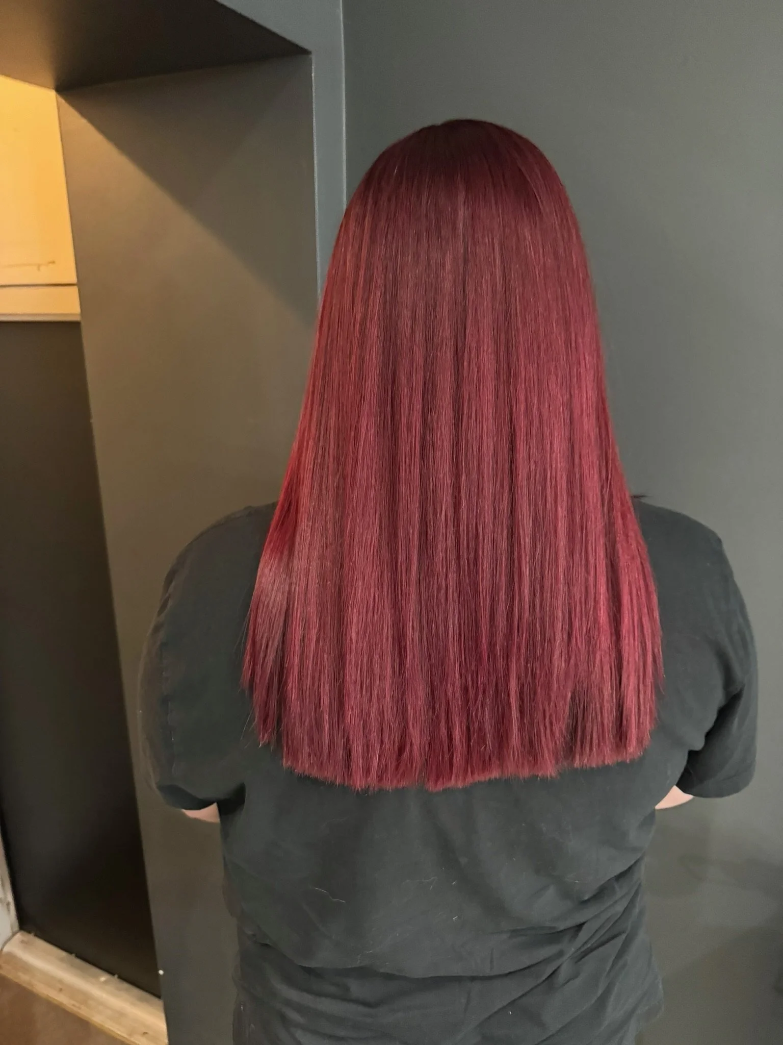 Back view of a person with long, straight, vibrant red hair wearing a dark shirt, standing indoors.